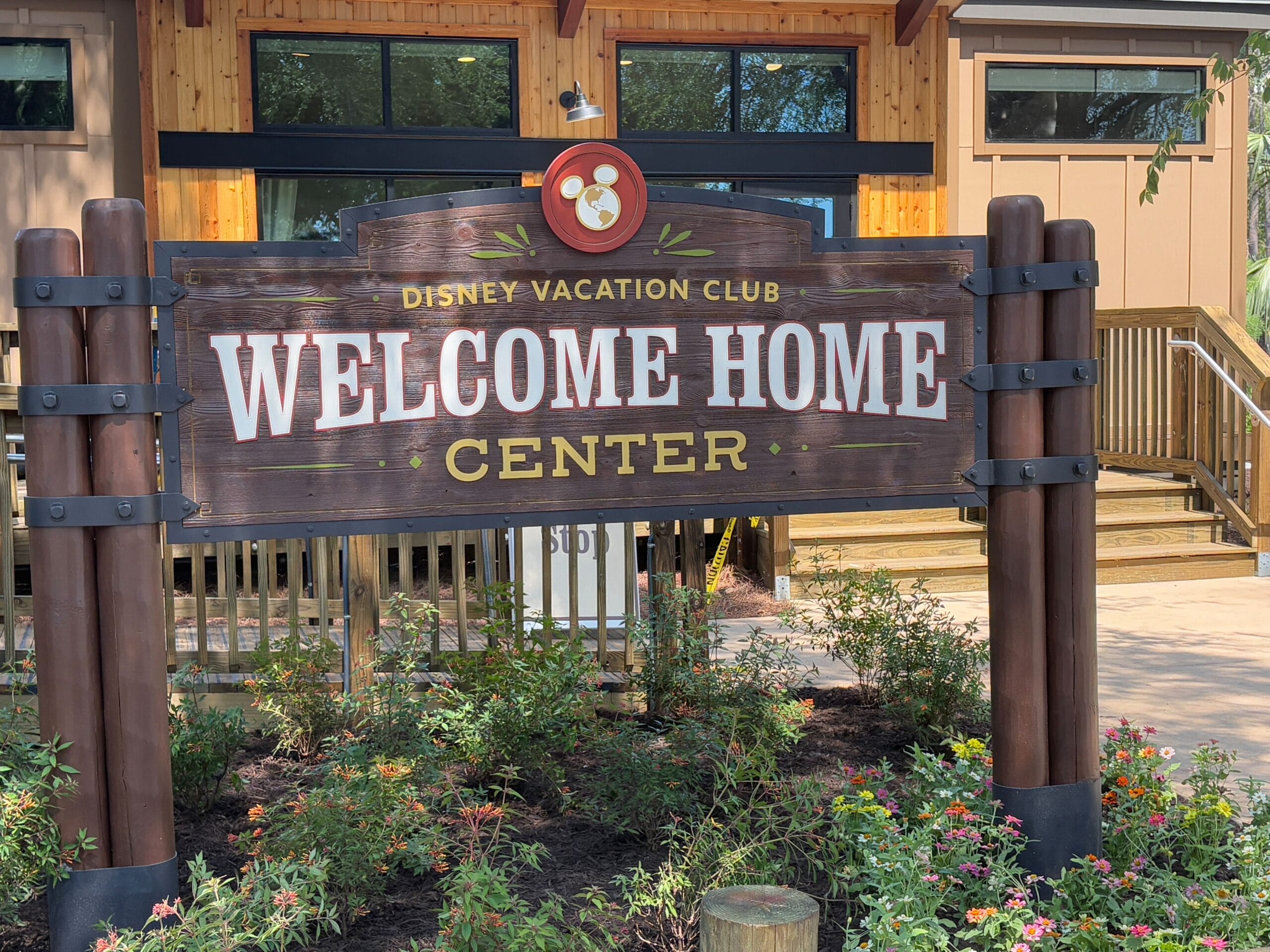 Wooden sign reading "Disney Vacation Club Welcome Home Center," surrounded by flowers and plants in front of a building.