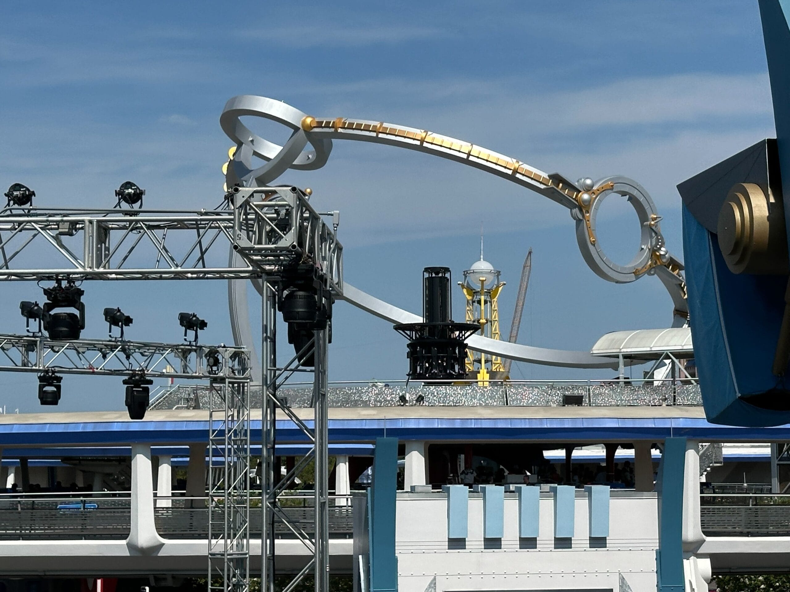 Metallic stage lights and sci-fi structures near the Astro Orbiter by a blue and white building under a clear sky.