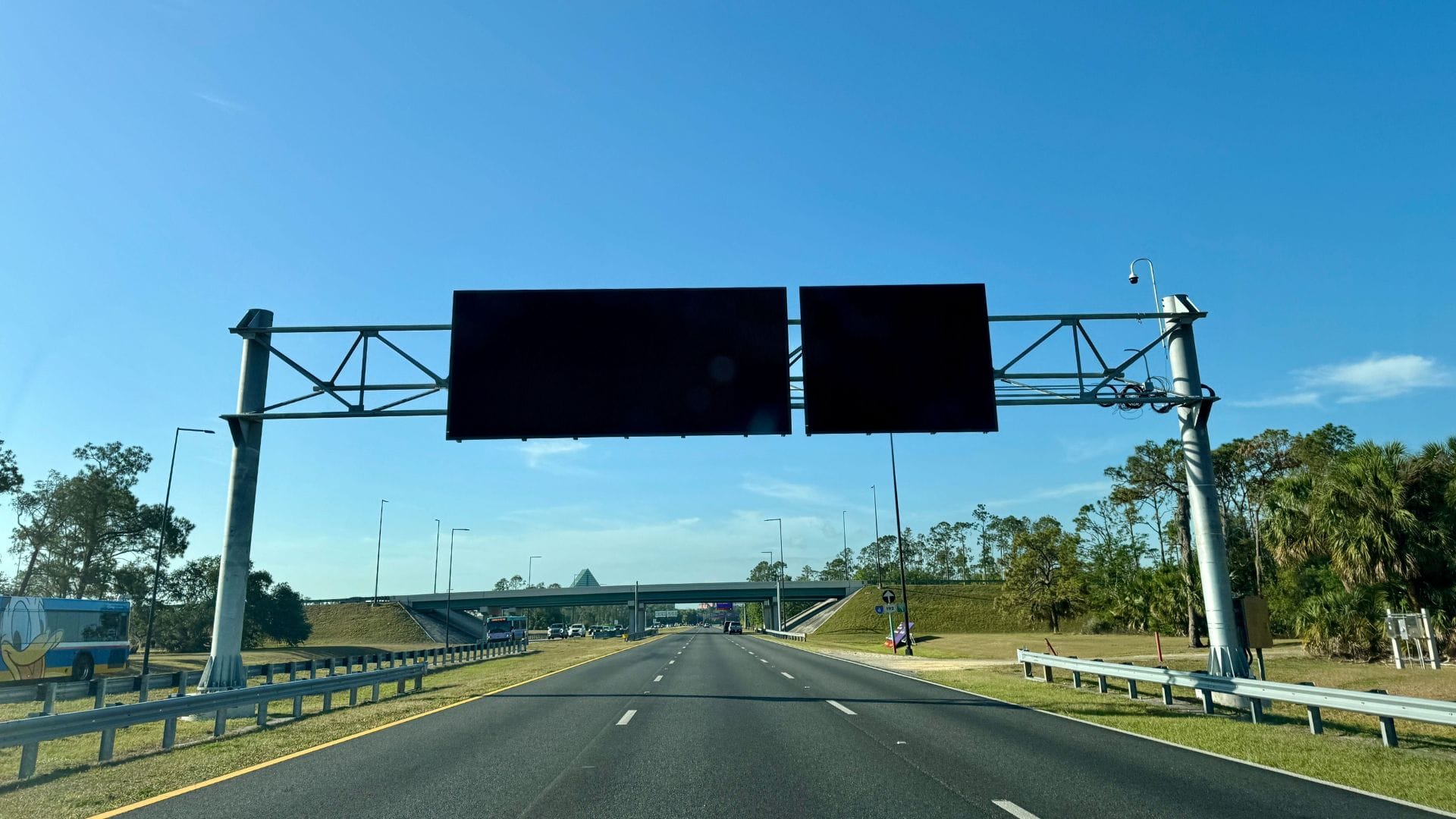 Two blank electronic highway signs above a clear multi-lane road on a sunny day with blue sky and trees.