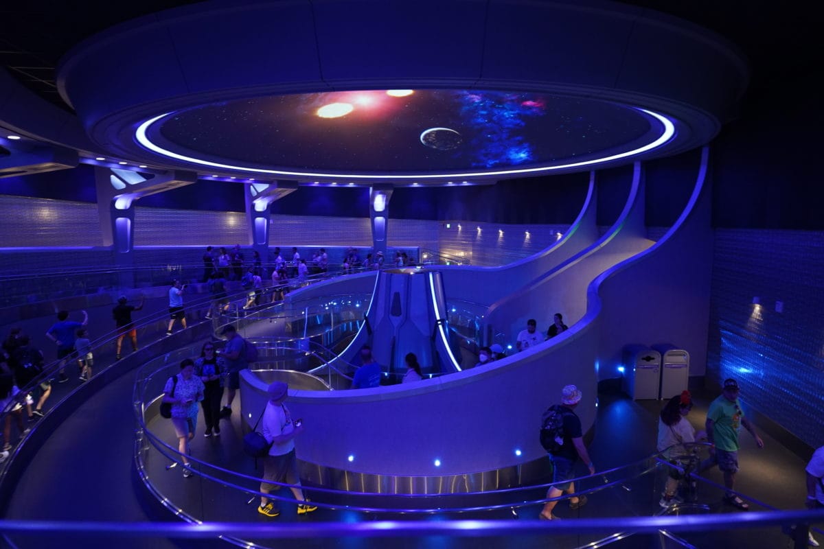 People walk along a futuristic, blue-lit ramp toward the Guardians of the Galaxy Ride at this space-themed attraction.