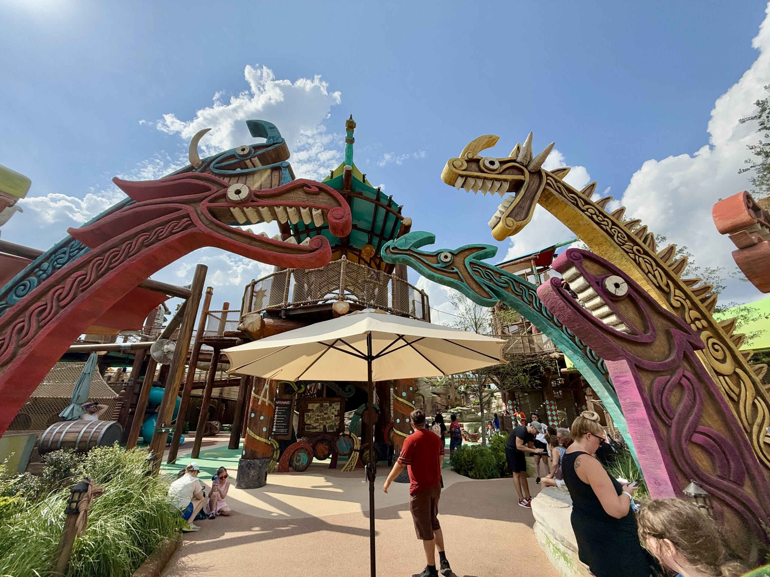 Colorful dragon sculptures arch over Epic Universe’s Viking Training Camp, where people play under a sunny sky.