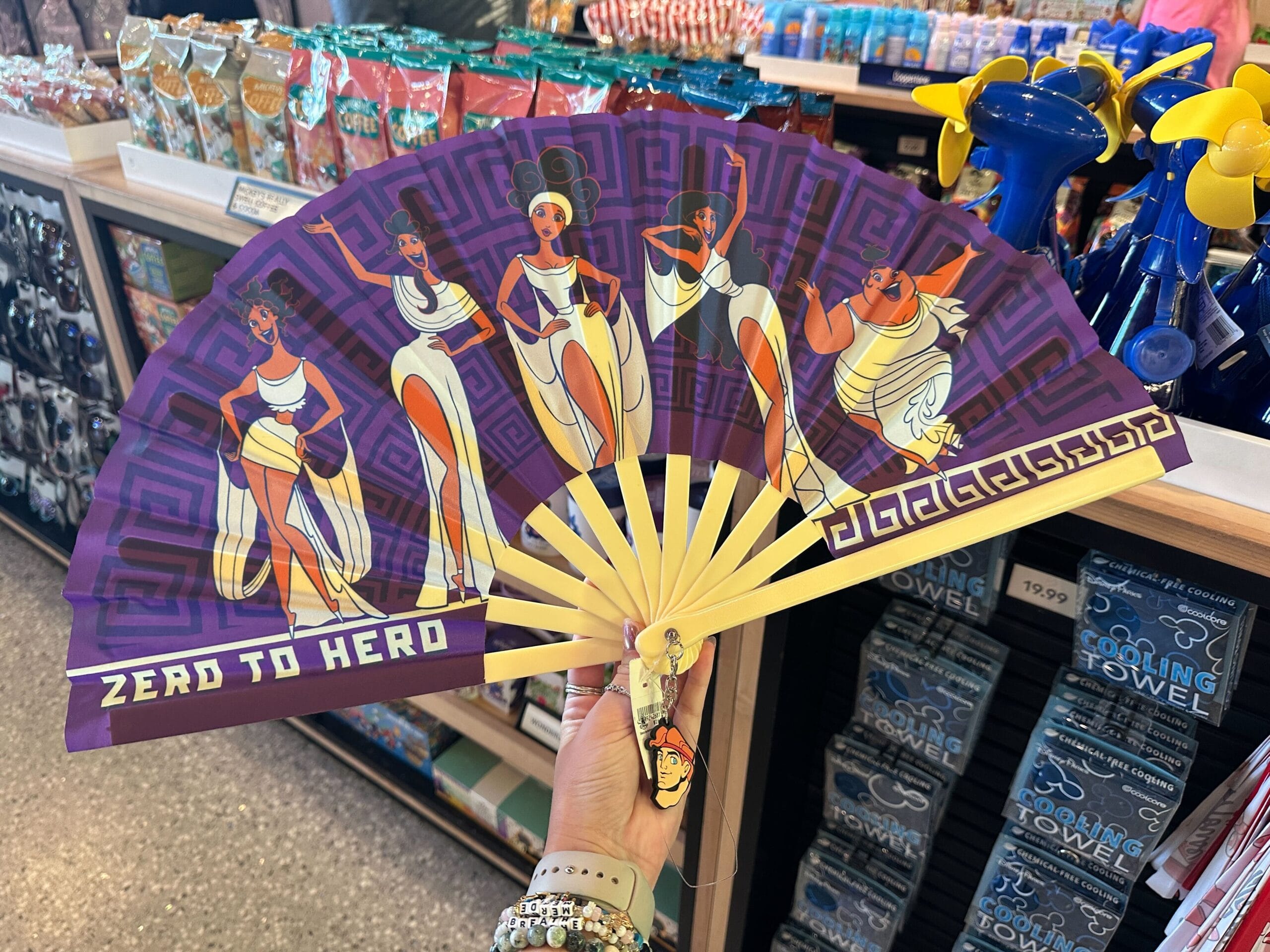 A hand displays a New Hercules Muse Hand Fan with “Zero to Hero” text in a Disney World store.
