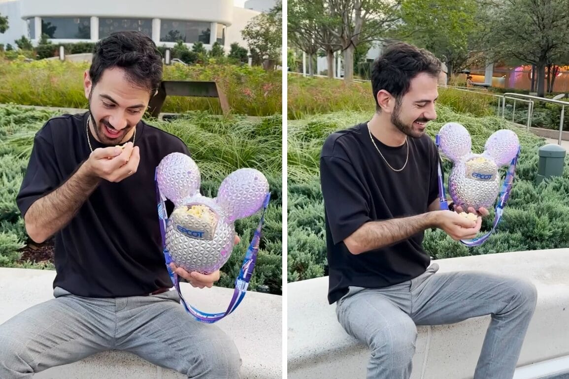 A man smiles while eating popcorn from a Mickey Balloon Popcorn Bucket outside in a park setting.