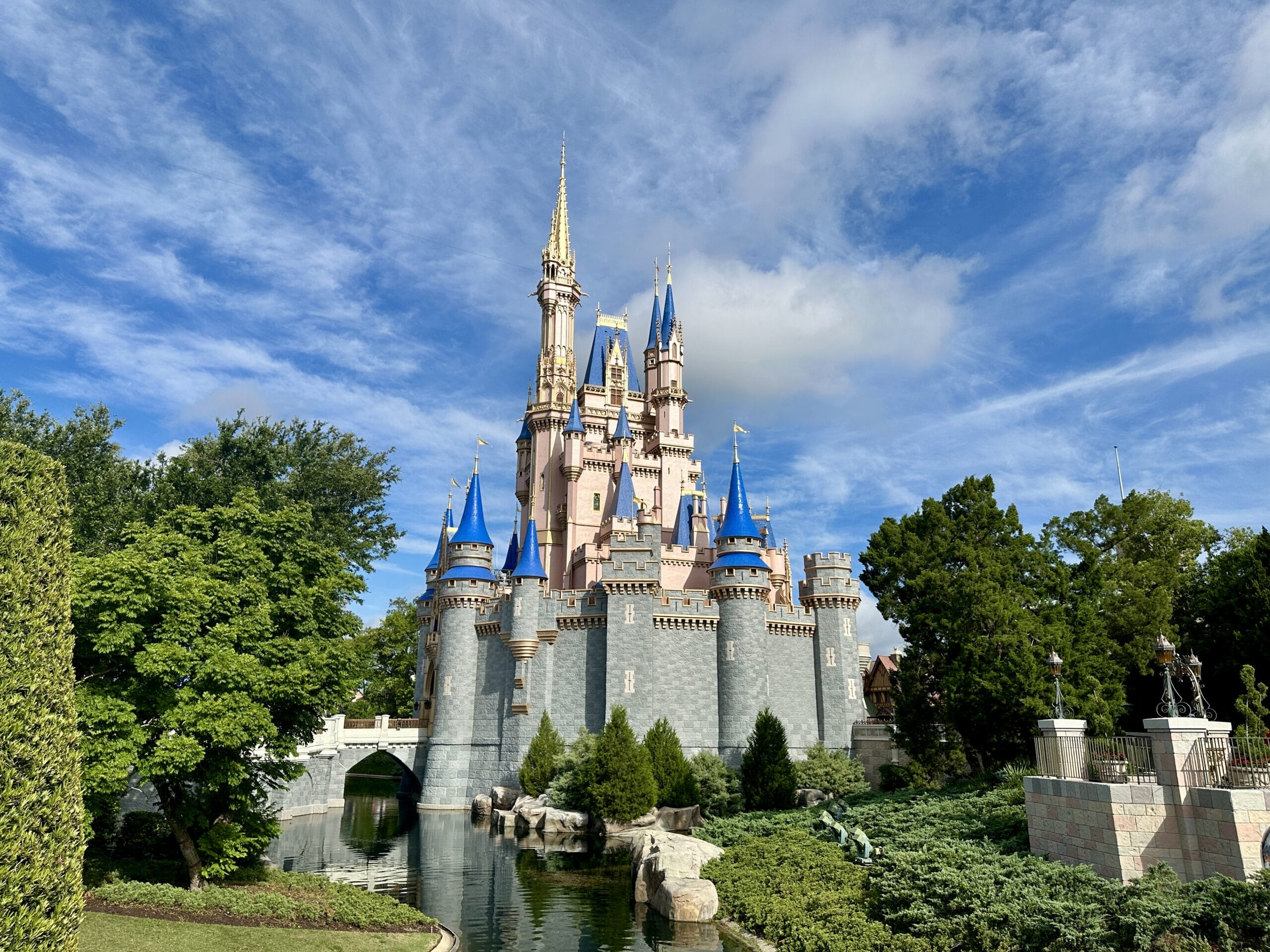 A fairytale castle with blue spires sits by a moat in Magic Kingdom, surrounded by green trees and a partly cloudy sky.
