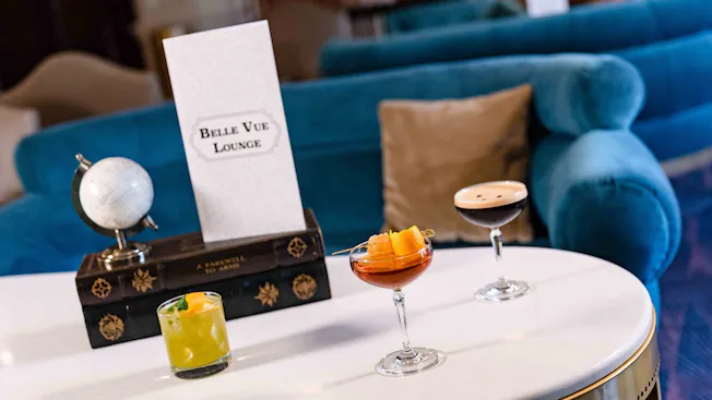 Three colorful cocktails on a white table in the chic Belle Vue Lounge with blue sofas and a signature lounge sign.
