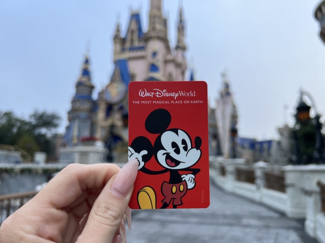 A hand holds a red Walt Disney World card with Mickey Mouse, ready to park hop Disney World by Cinderella’s Castle.