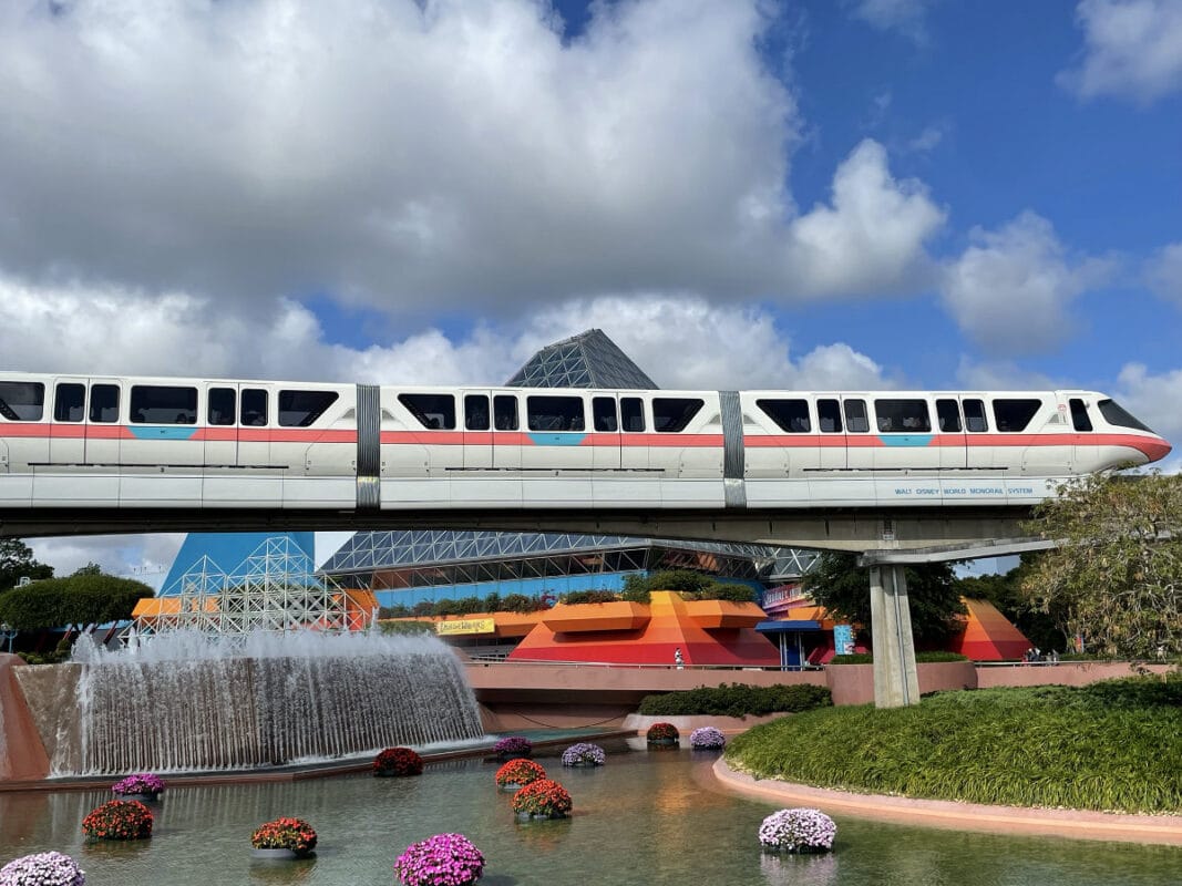 A white and red monorail glides above a fountain at Disney World, with colorful buildings under a partly cloudy sky.