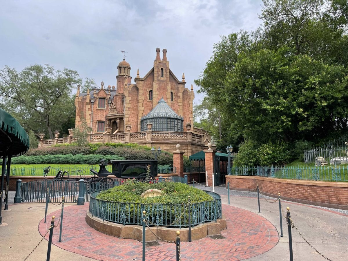 magic kingdom haunted mansion entrance with gothic architecture sits behind a circular garden and wrought iron fence on a cloudy day.