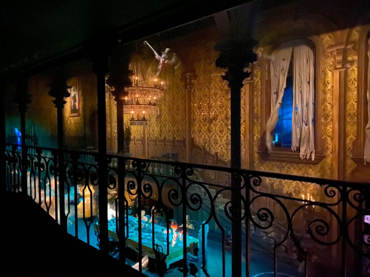 the ballroom of the haunted Mansion in disney world magic kingdom with chandeliers, draped windows, and a balcony overlooking a dining table.