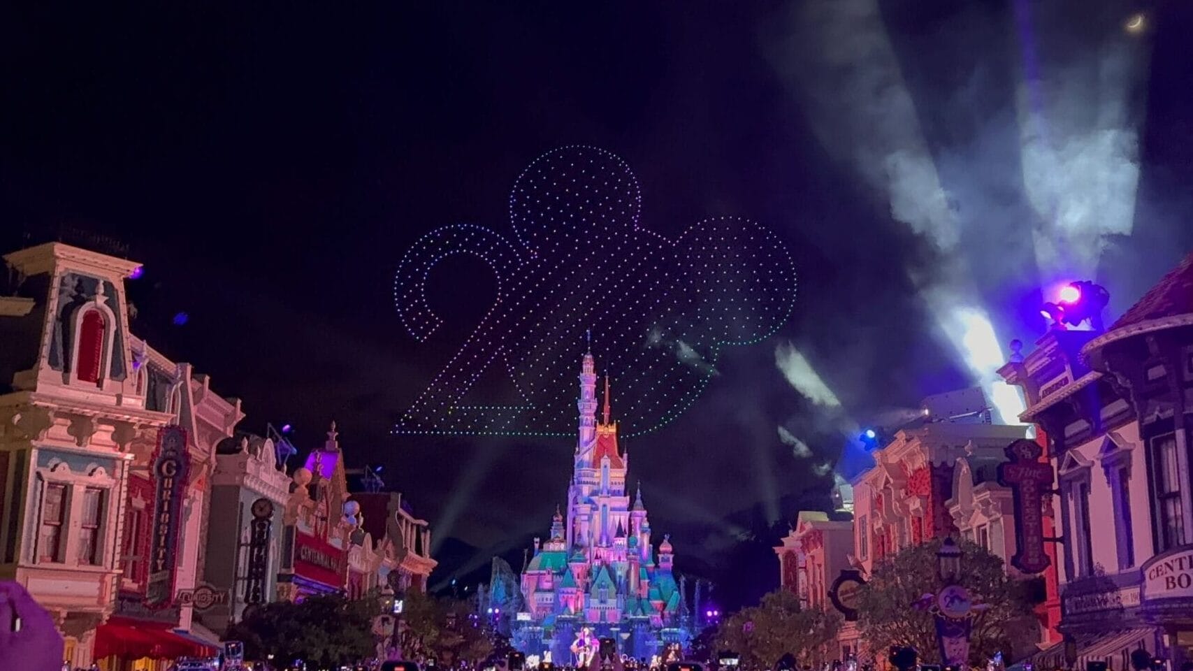 A nighttime crowd faces a lit-up castle during the 20th Anniversary Drone Pre-Show and "Disney 100" sky display.