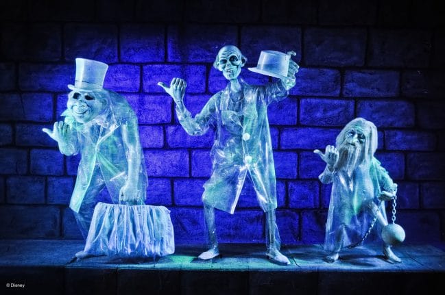 the hitchhiking ghotst in suits and hats, glowing blue, stand by a dark Haunted Mansion stone wall.