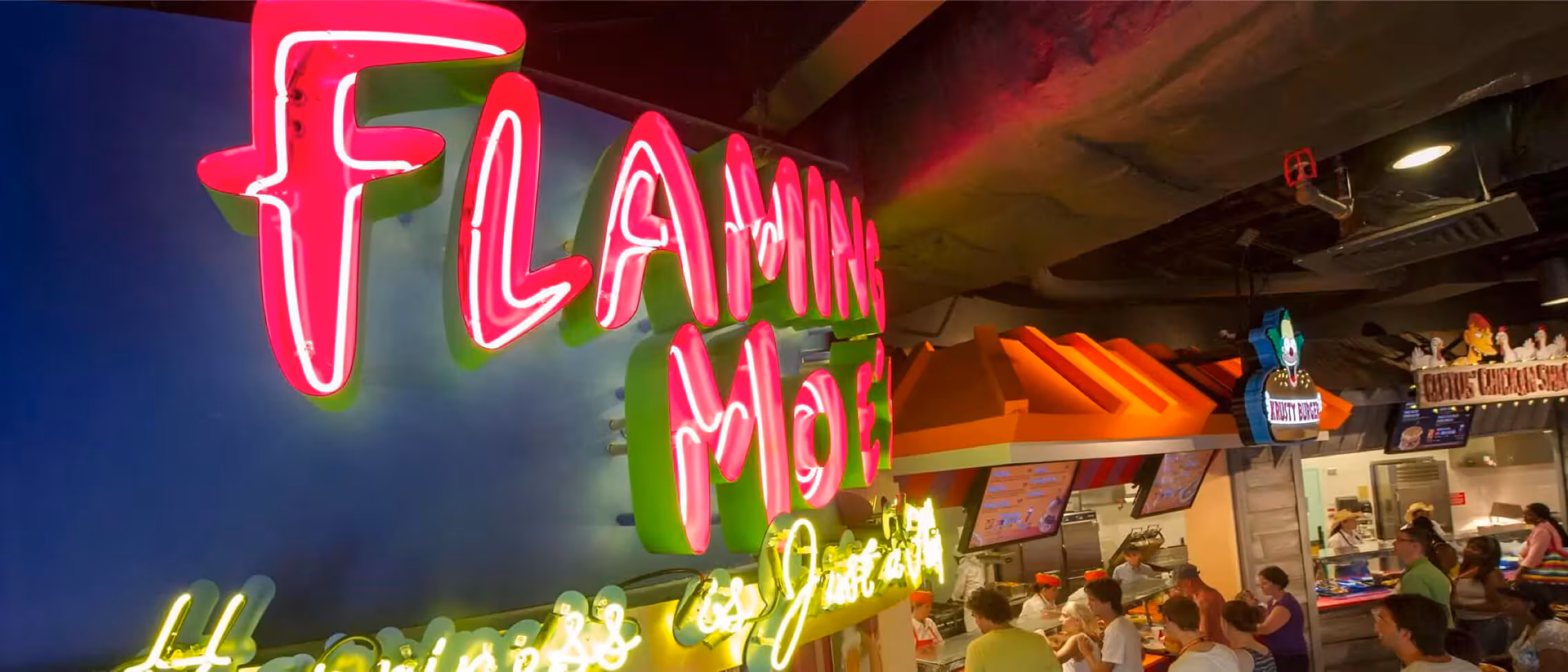 Neon "Flaming Moe's" sign at a lively Simpsons-themed bar with people dining and ordering food at the counter.