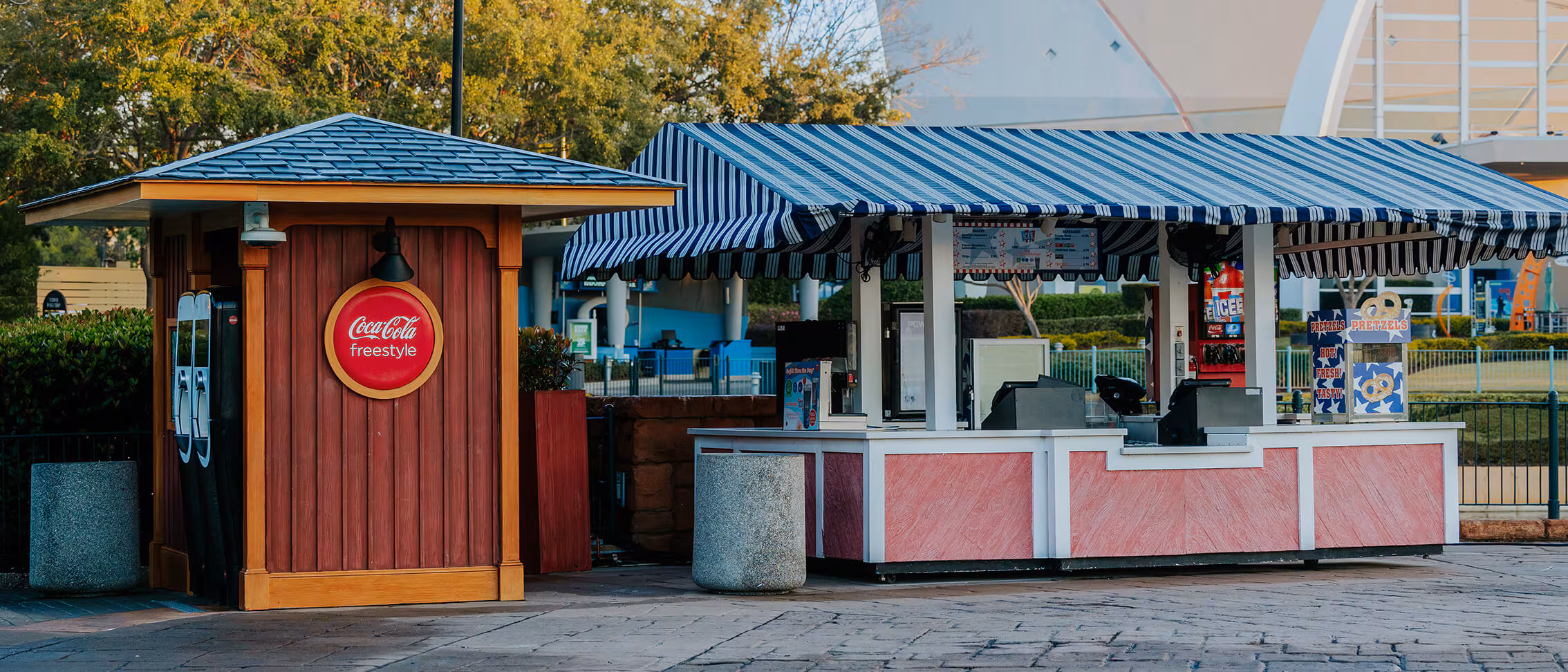 A small red Coca-Cola Freestyle booth offers Lagoon Lemonade by a striped-roof concession stand in an outdoor park.