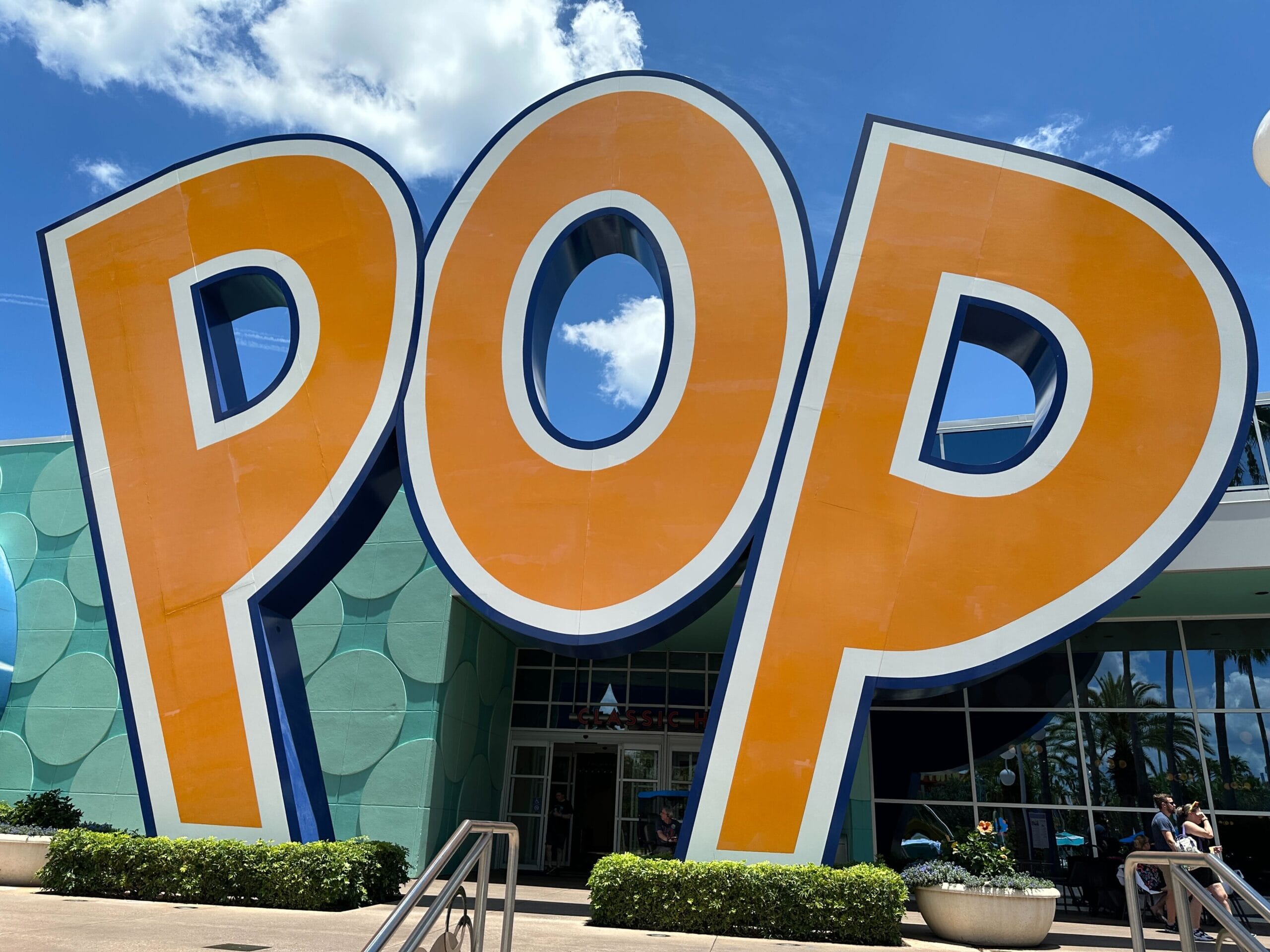 Large orange letters spelling "POP" stand in front of a colorful building under a blue sky with clouds.