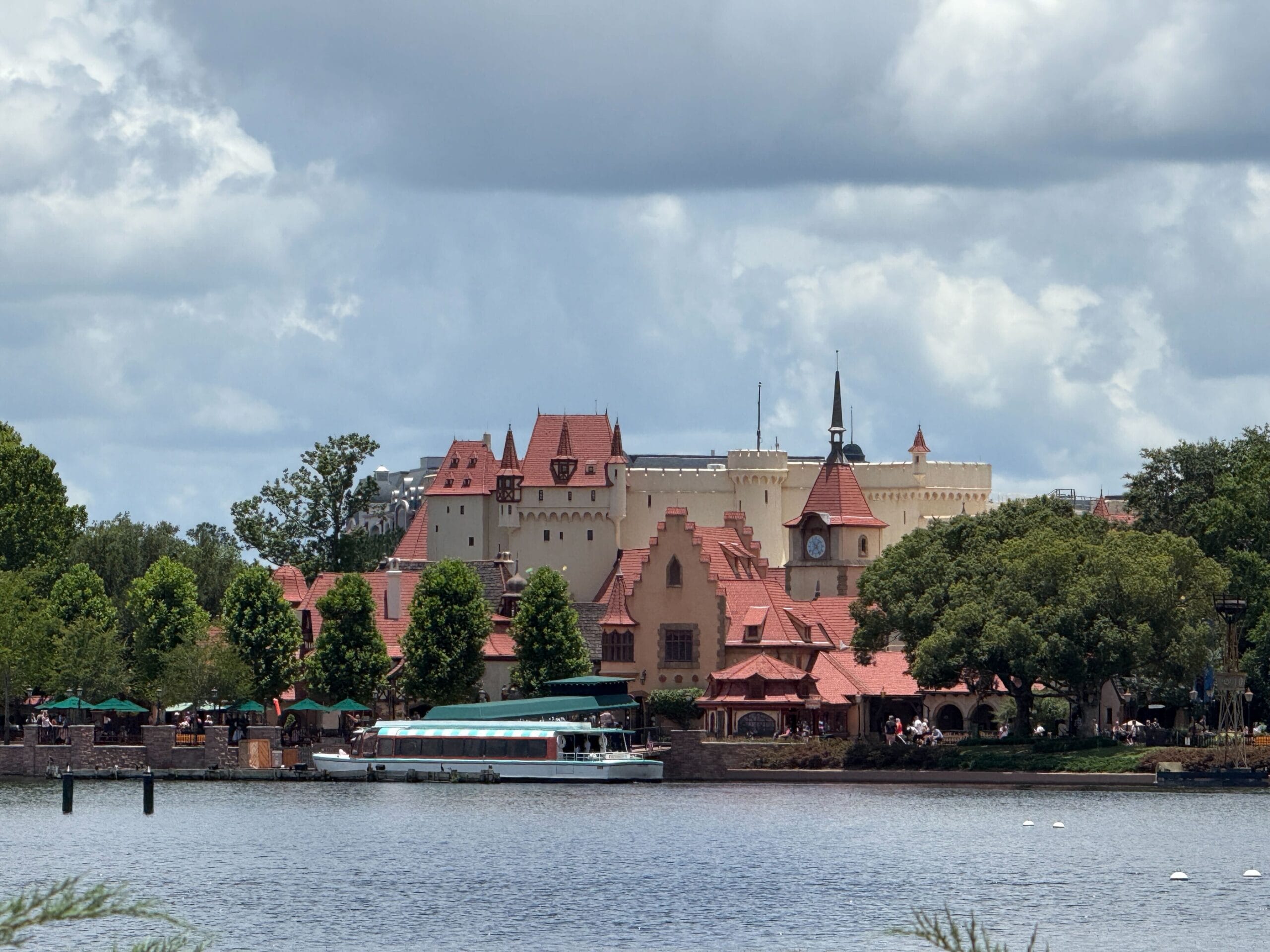 A Friendship Boat passes a village with red roofs and clock tower at EPCOT World Showcase under a cloudy sky.