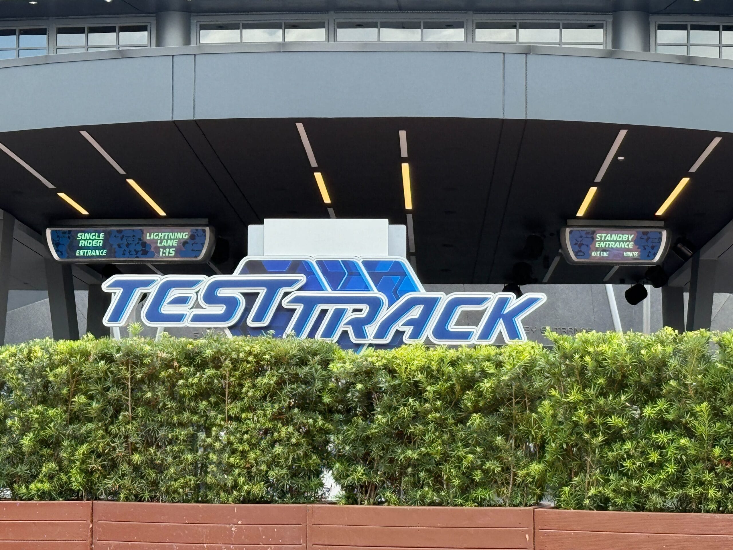 Entrance to Test Track ride with digital wait time signs behind greenery at a theme park.