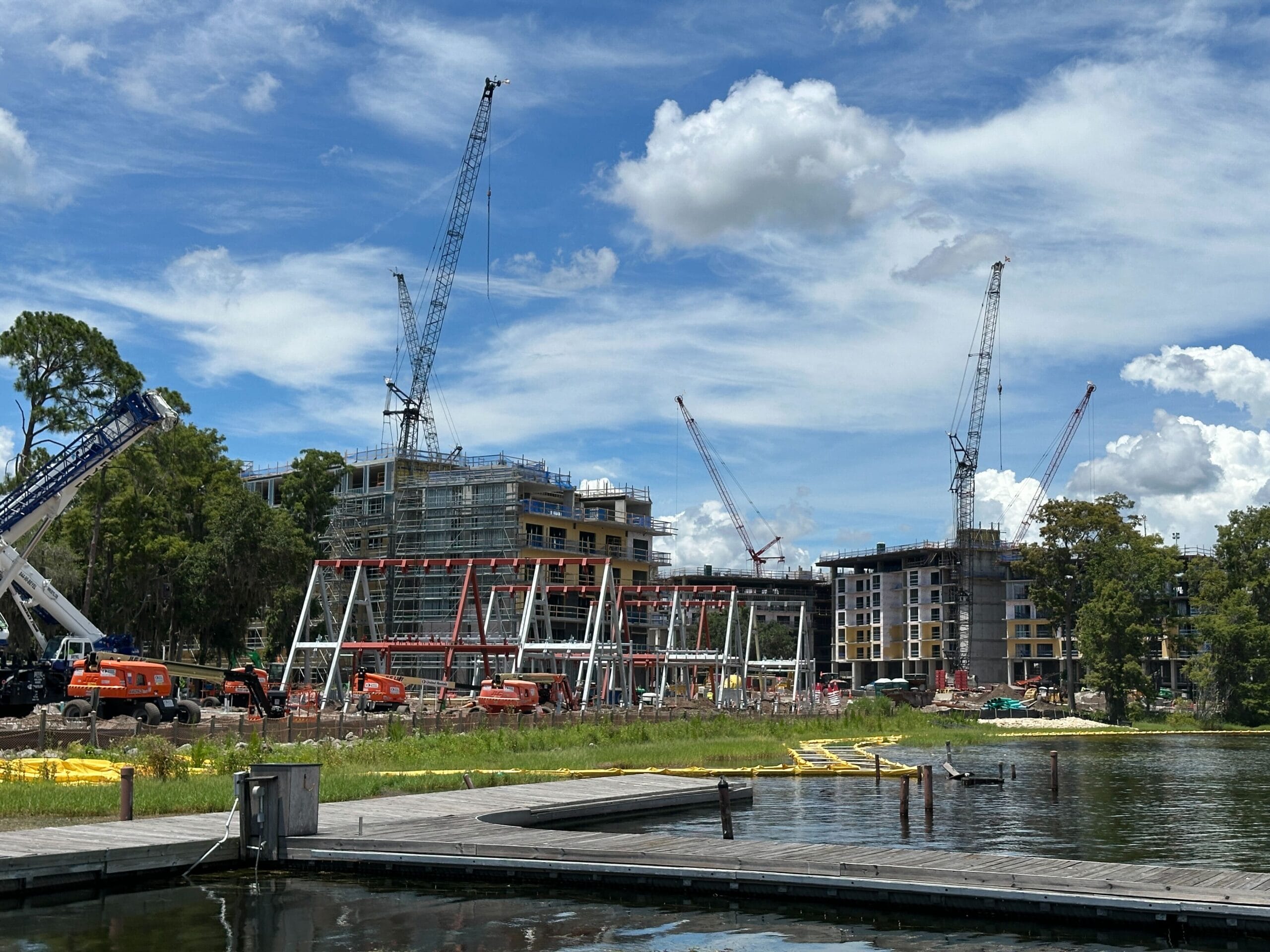 Construction cranes and equipment work on buildings near a lake under a partly cloudy sky.