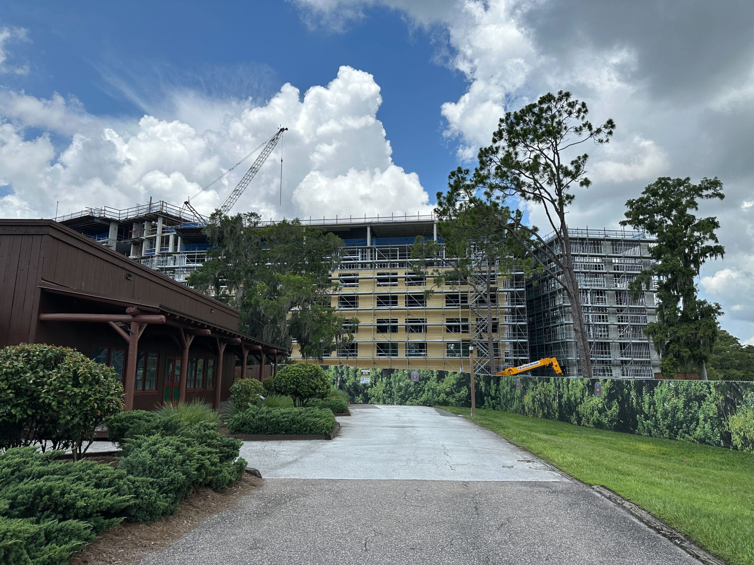 A multi-story Lakeshore Lodge under construction with scaffolding, crane, and cloudy sky, behind a green fence.