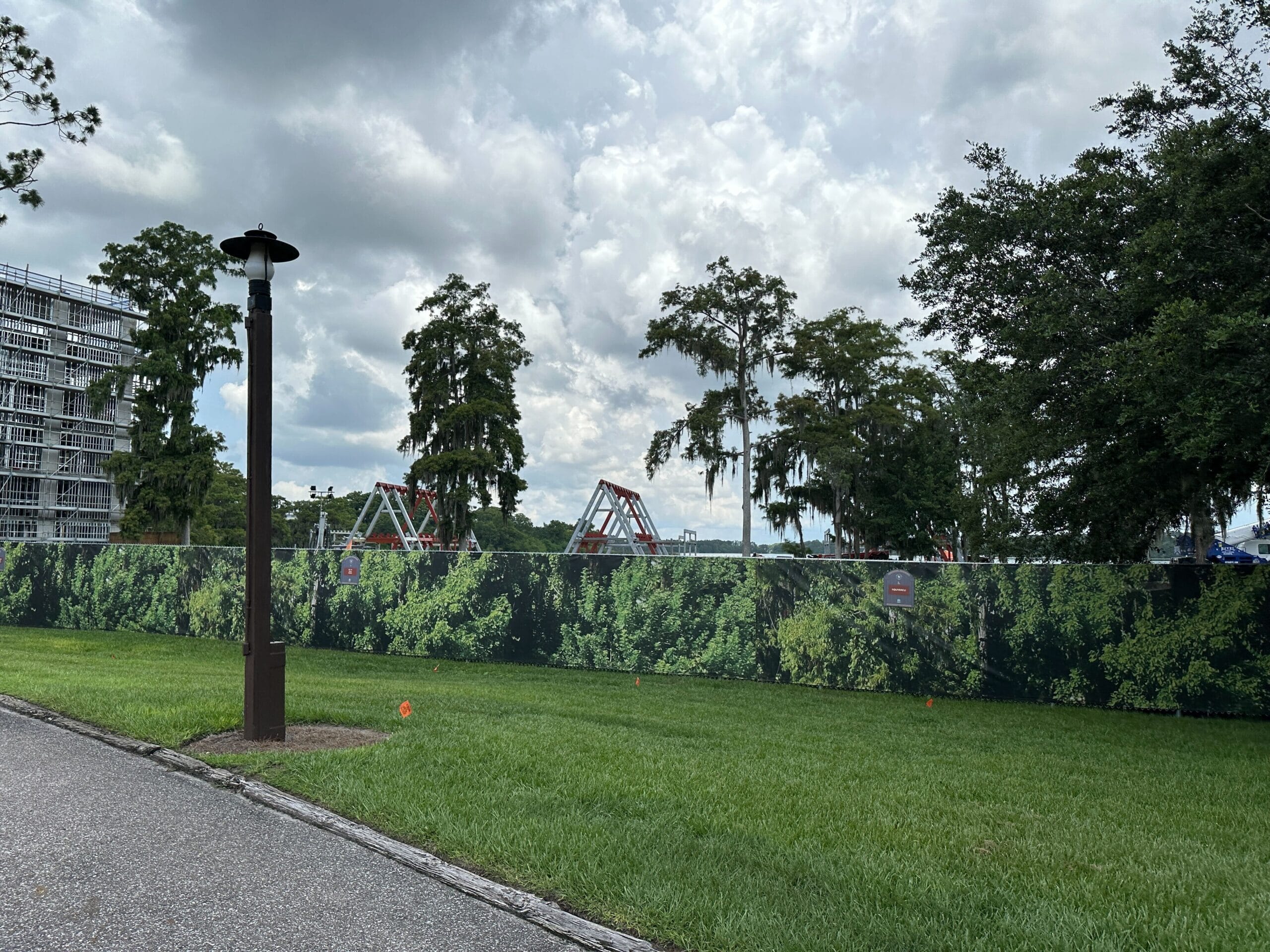 A construction site for Lakeshore Lodge behind a leafy fence, with trees, grass, and a cloudy sky in the background.