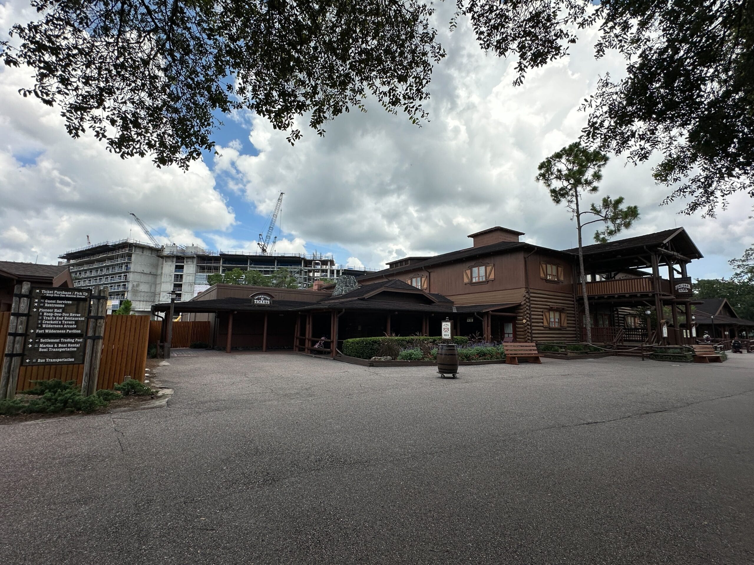 A rustic Lakeshore Lodge with a gravel lot, trees, and cranes behind under a cloudy sky.