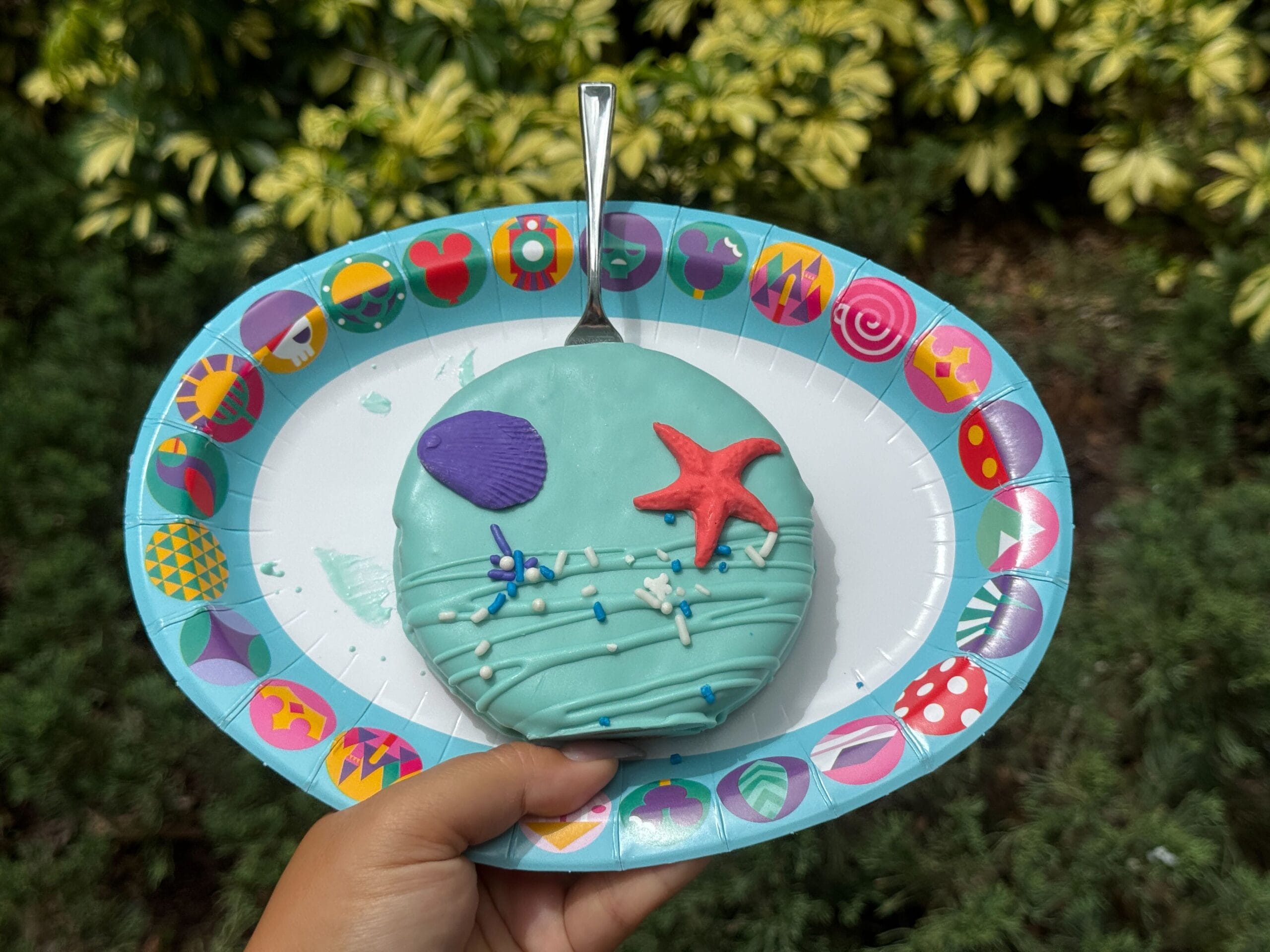 A hand holds a blue Little Mermaid Cookie Pie cake with a starfish, outdoors on a colorful plate amid green scenery.
