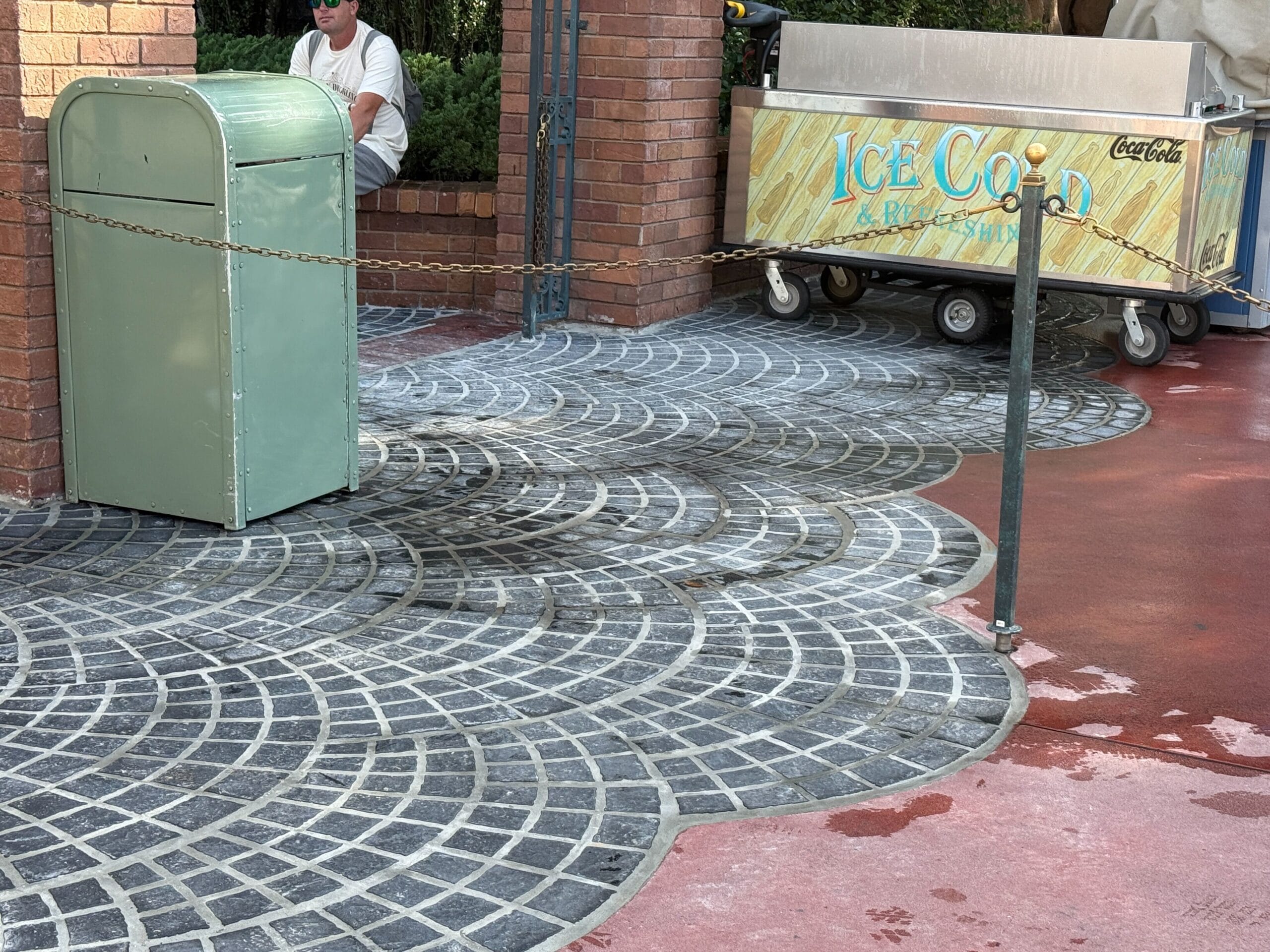 A green trash can, an ice cream cart, and a man sitting on a bench near patterned pavement and red concrete.