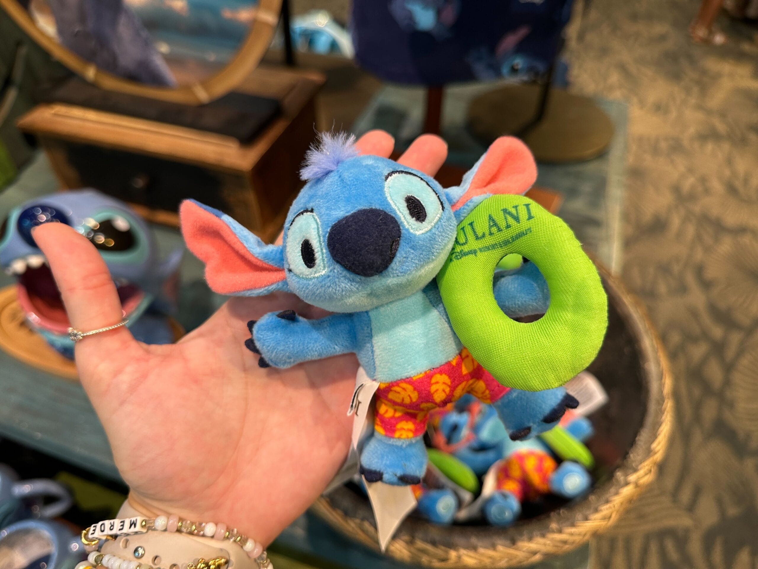 A hand holds a small Stitch plush toy wearing red shorts and holding a green floatie labeled "AULANI.