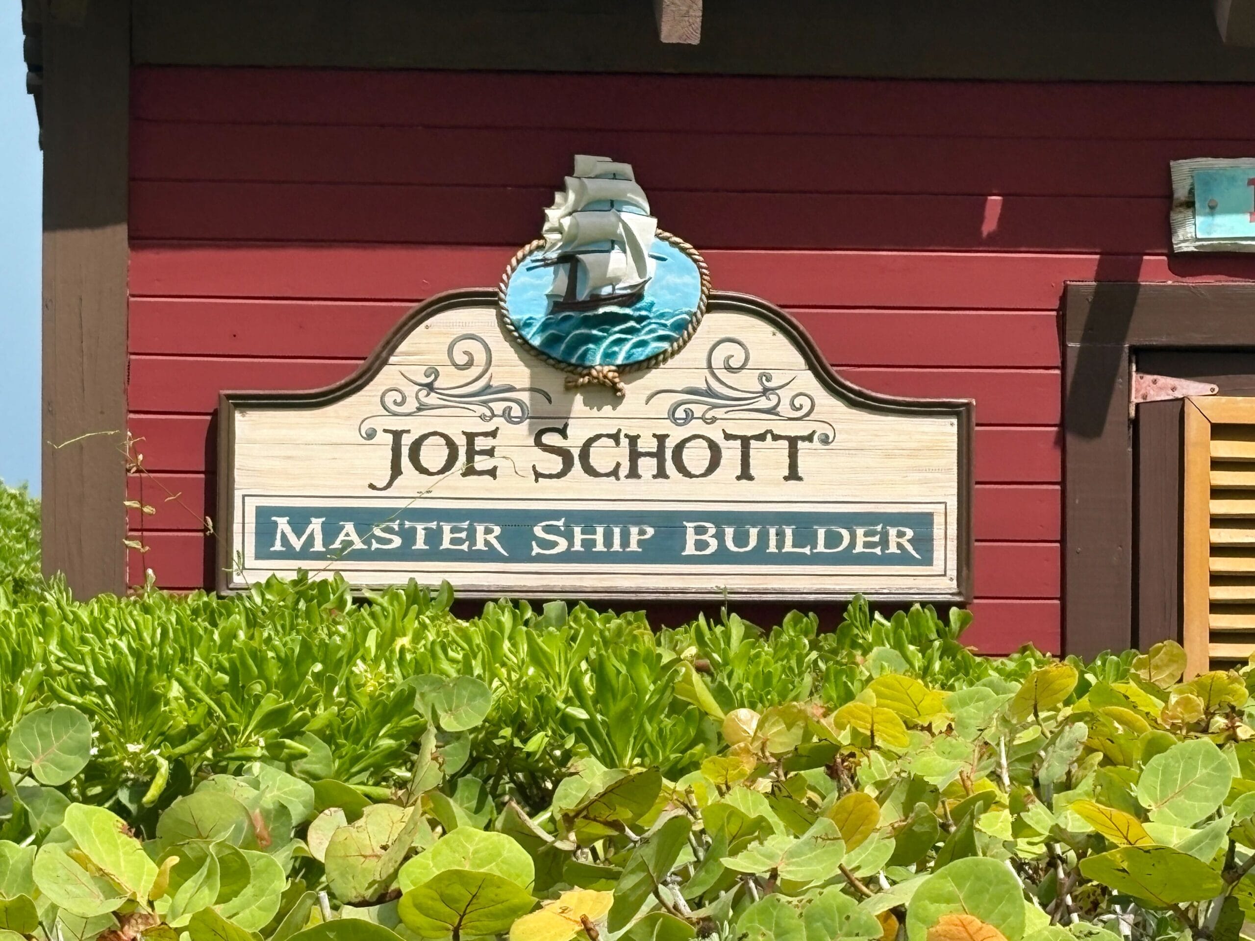 A sign reads "Joe Schott Master Ship Builder" with a ship emblem, set on Castaway Cay's red wooden building.