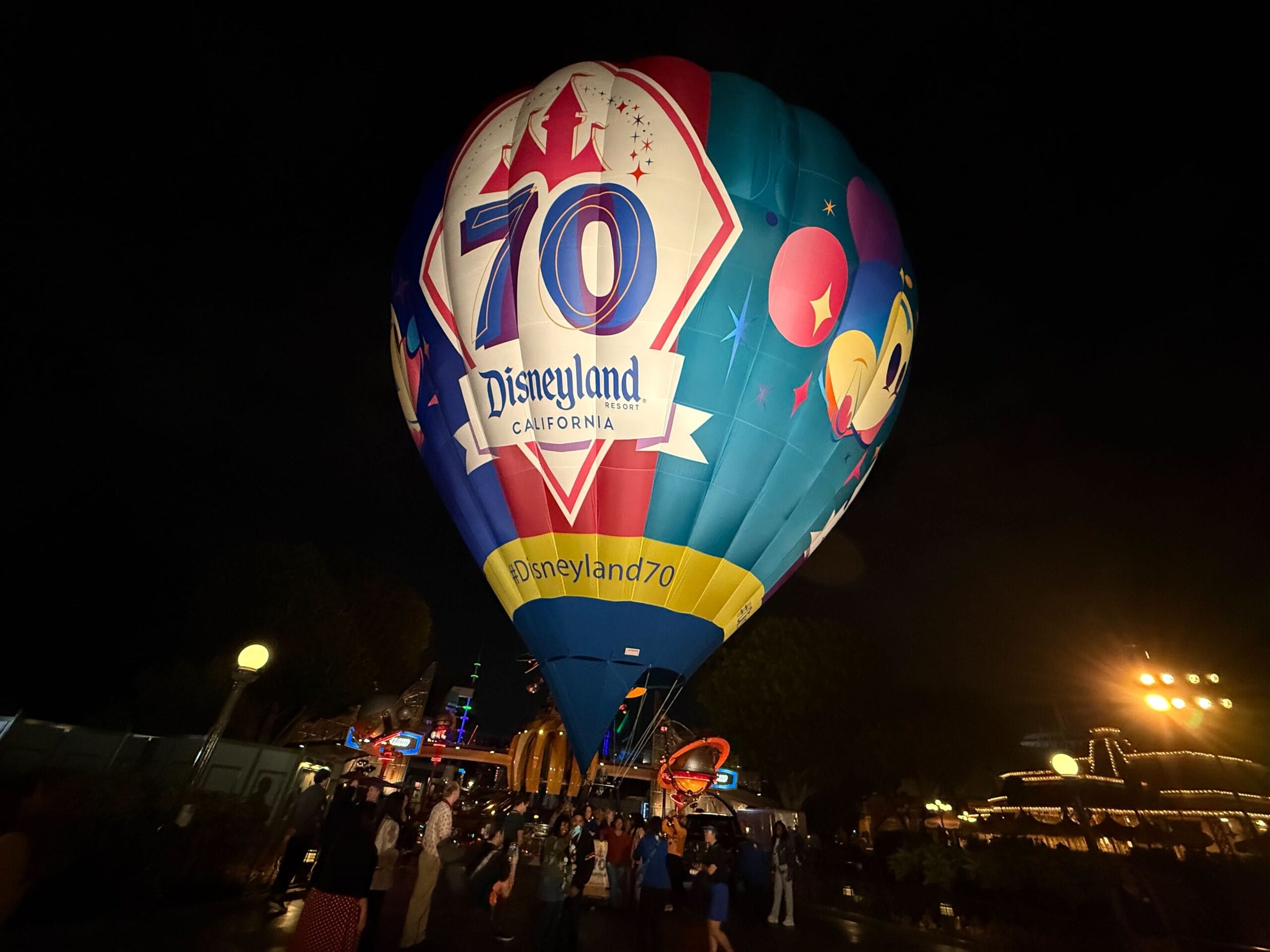 A Hot Air Balloon with a “70th Anniversary Disneyland” logo glows at night, drawing crowds amid sparkling park lights.