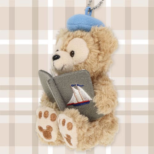 A plush Duffy The Disney Bear in a blue beret holds an open book with a sailboat, sitting against a plaid background.