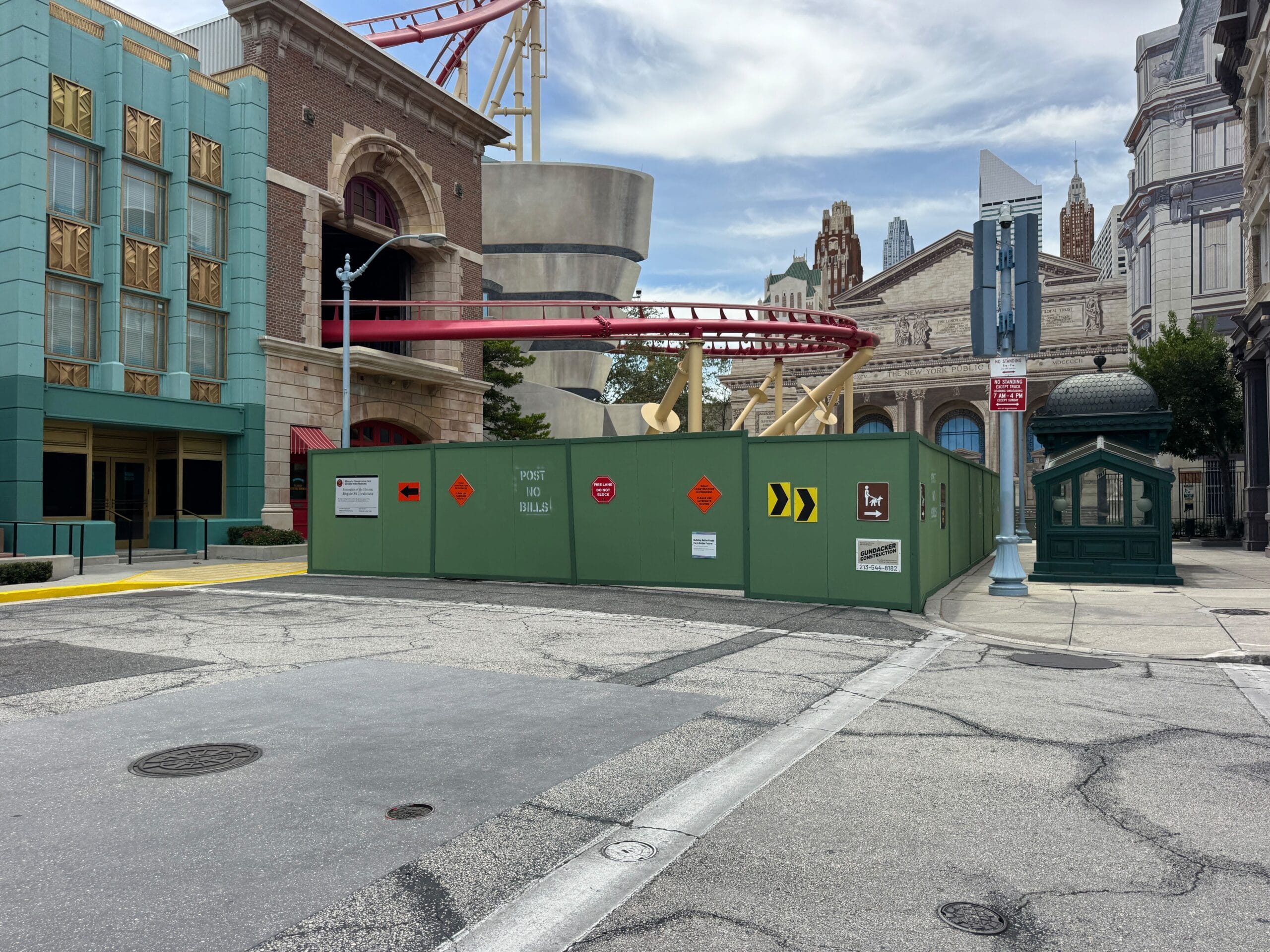 A theme park street with construction walls, warning signs, and a roller coaster track overhead under a partly cloudy sky.