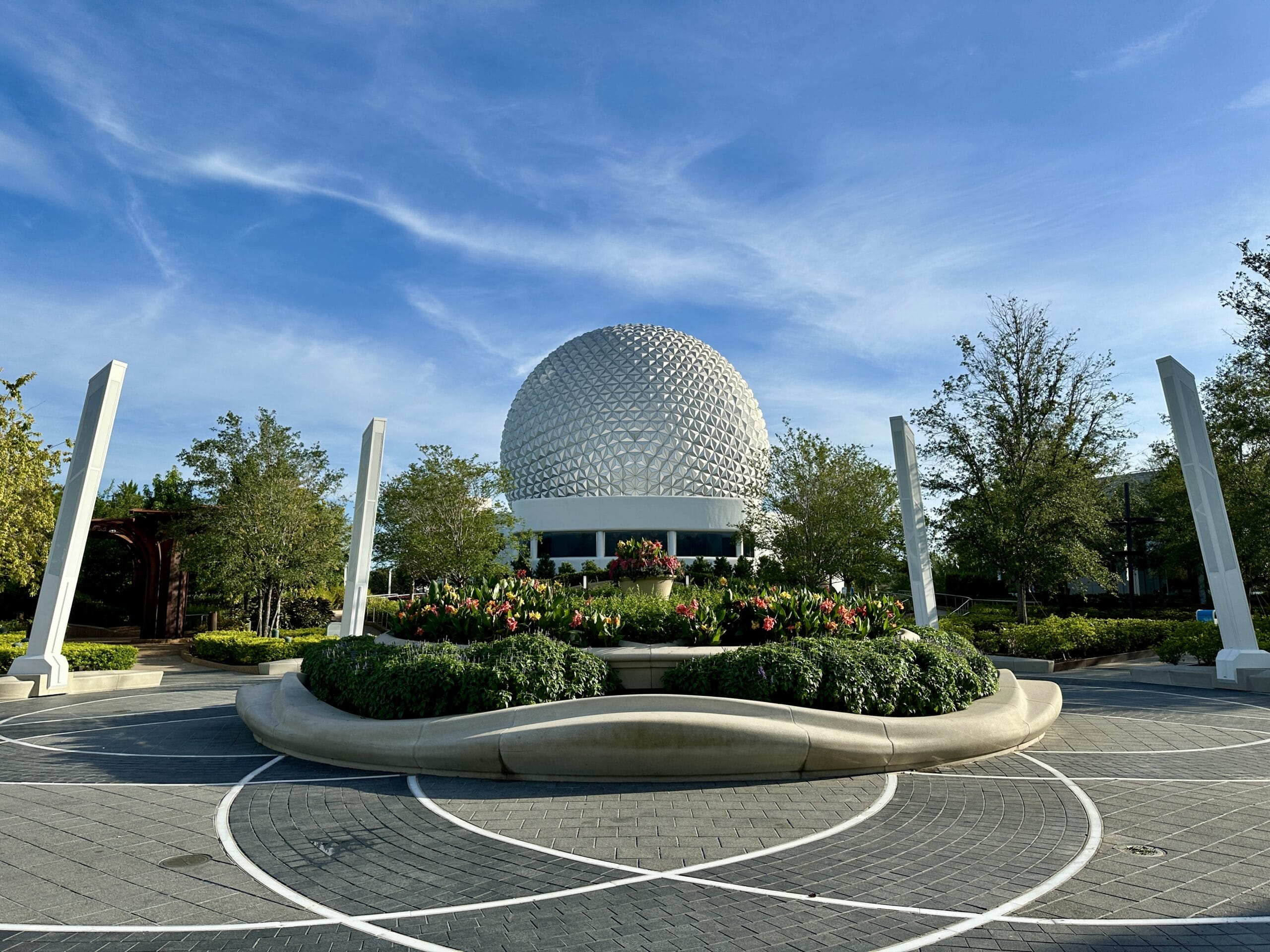 A large geodesic sphere at EPCOT stands behind a landscaped garden with flowers and trees under a blue sky.