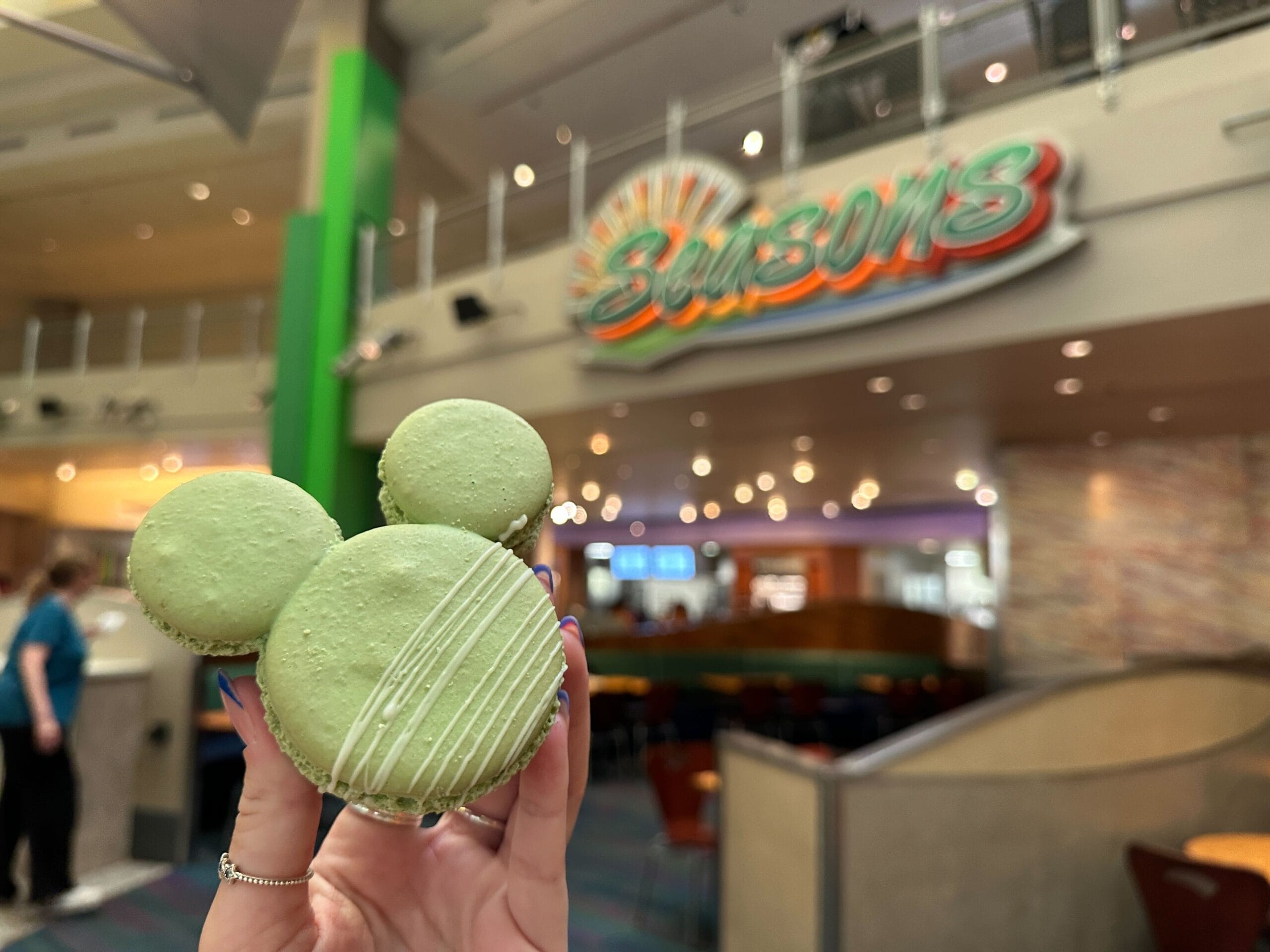 A hand holds a green Mickey-shaped macaron in front of a Seasons restaurant sign inside a bright building.