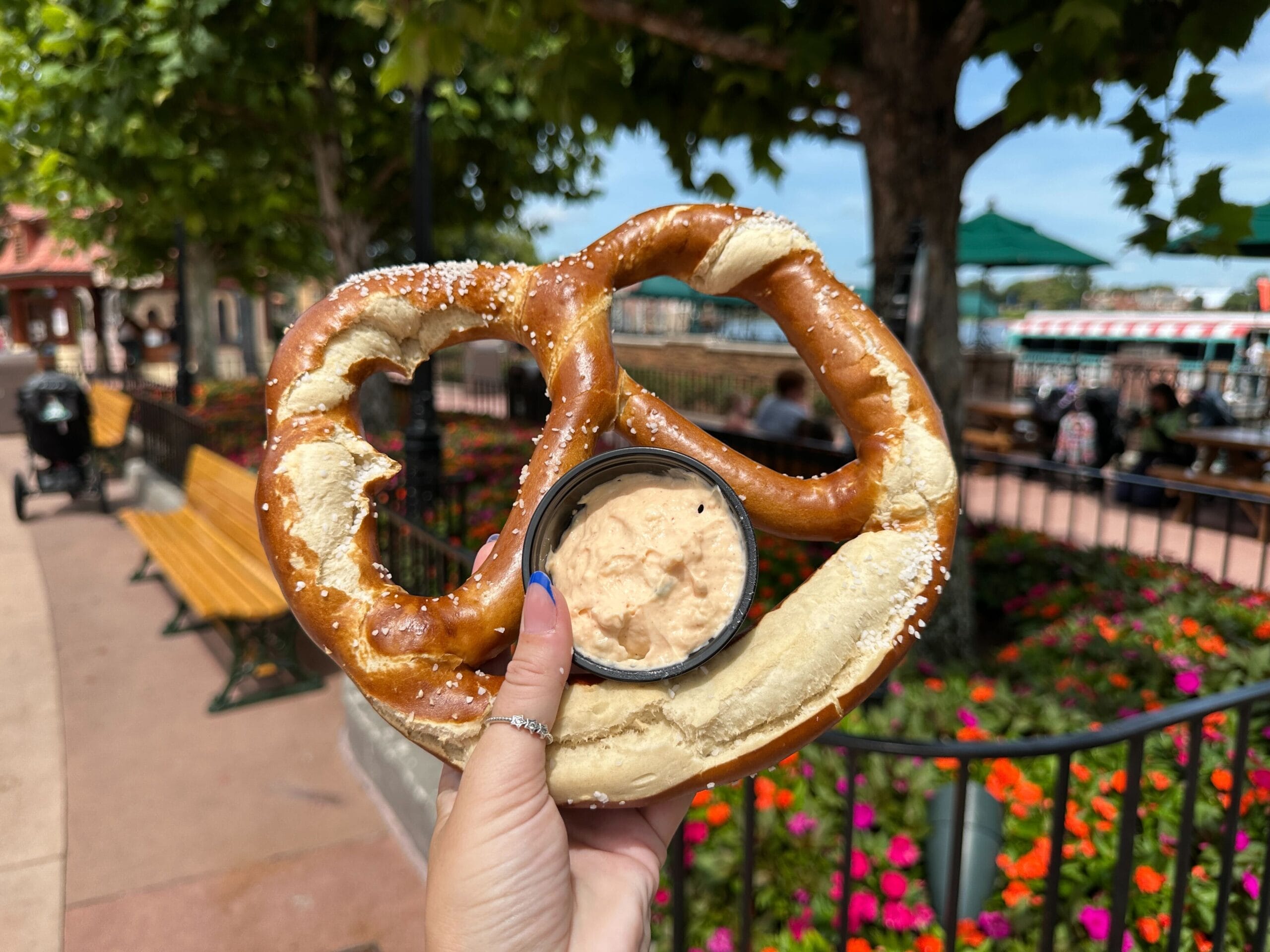 A hand holds a large soft pretzel with cheese dip, outdoors near benches and colorful flowers.