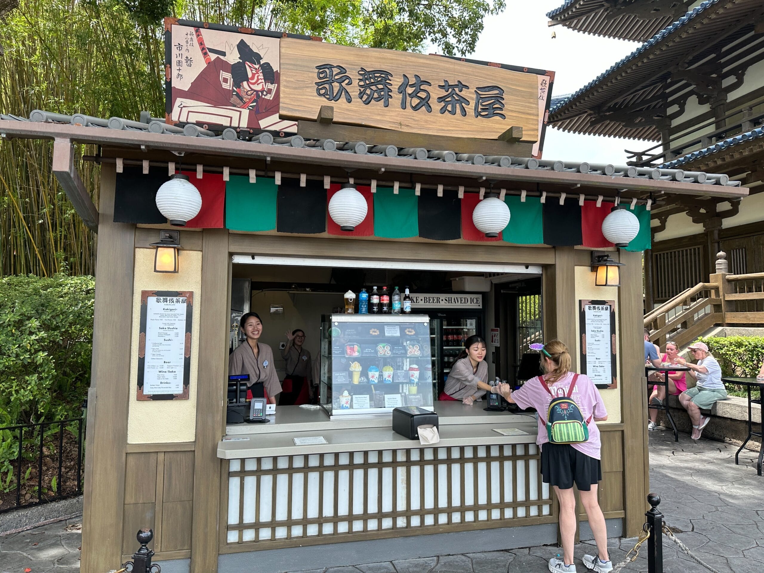 A Japanese tea and snack stand at EPCOT with staff serving a customer and colorful lanterns above the counter.