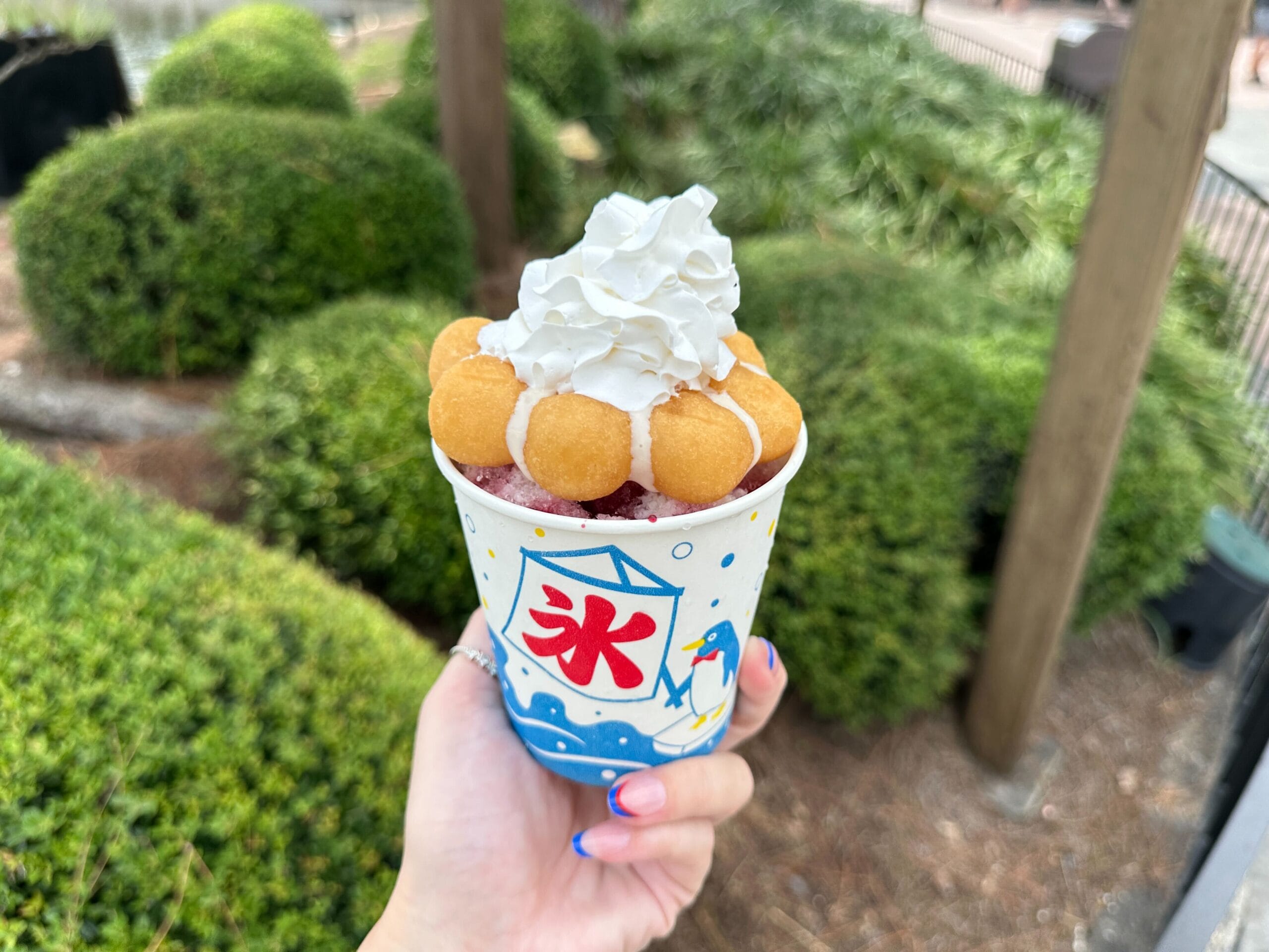 A hand holds a shaved ice dessert topped with cream and a Mickey-Shaped Pistachio Macaron at EPCOT, trees in the background.