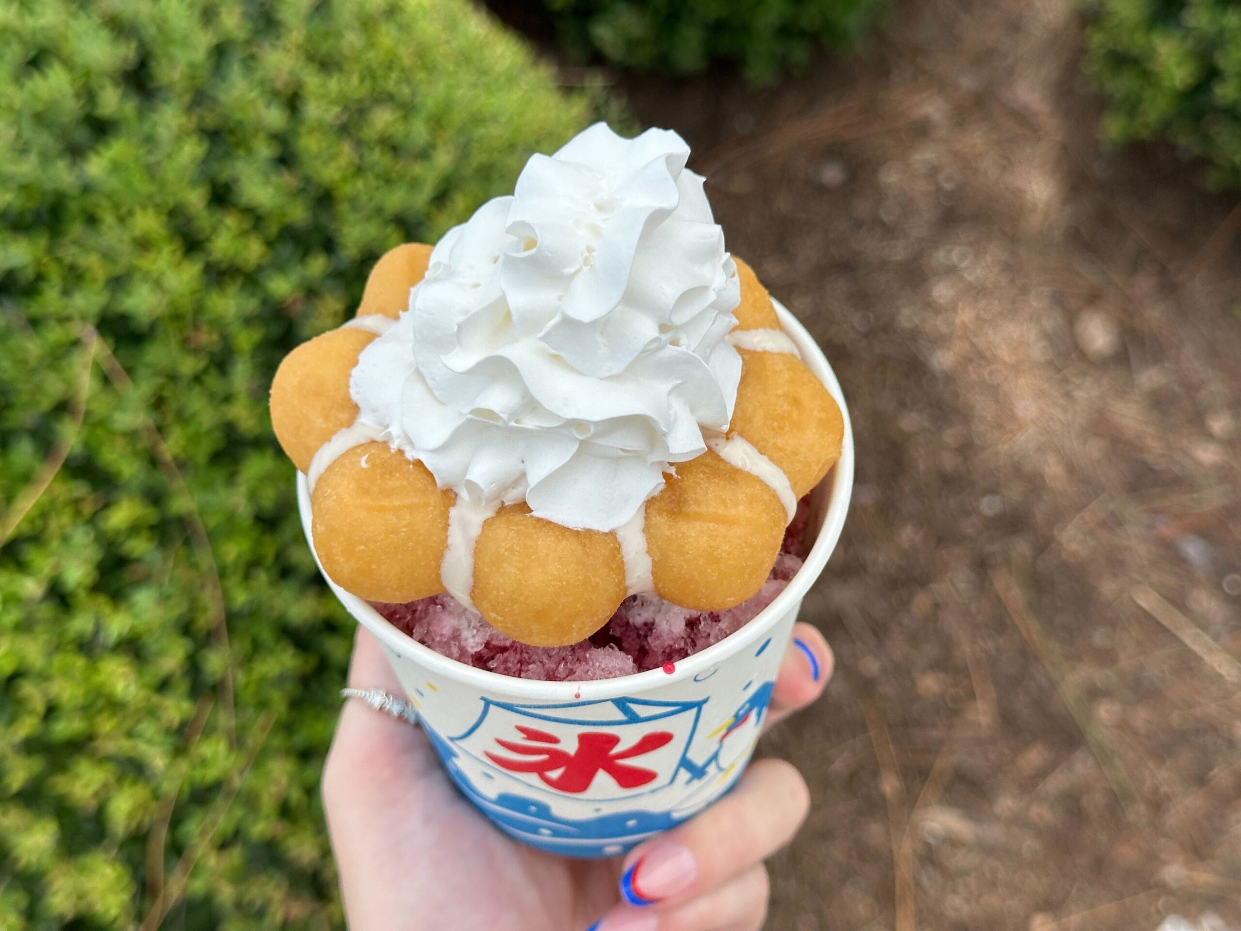 A hand holds a shaved ice dessert with whipped cream and pastry balls—one of the new EPCOT snacks to try.