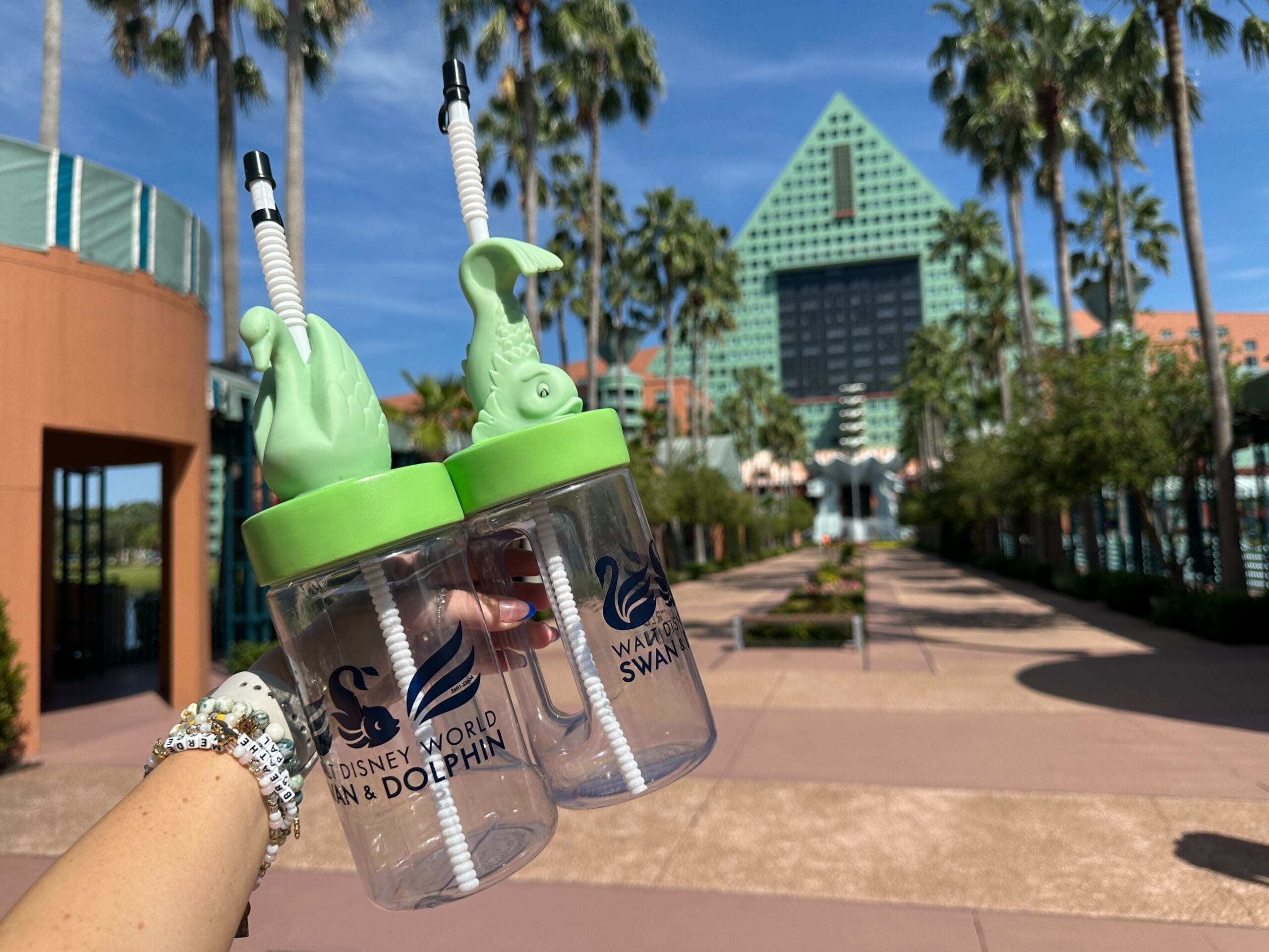 Two Swan and Dolphin Souvenir Sippers with dolphin lids are held up outside the resort under a sunny blue Walt Disney World sky.