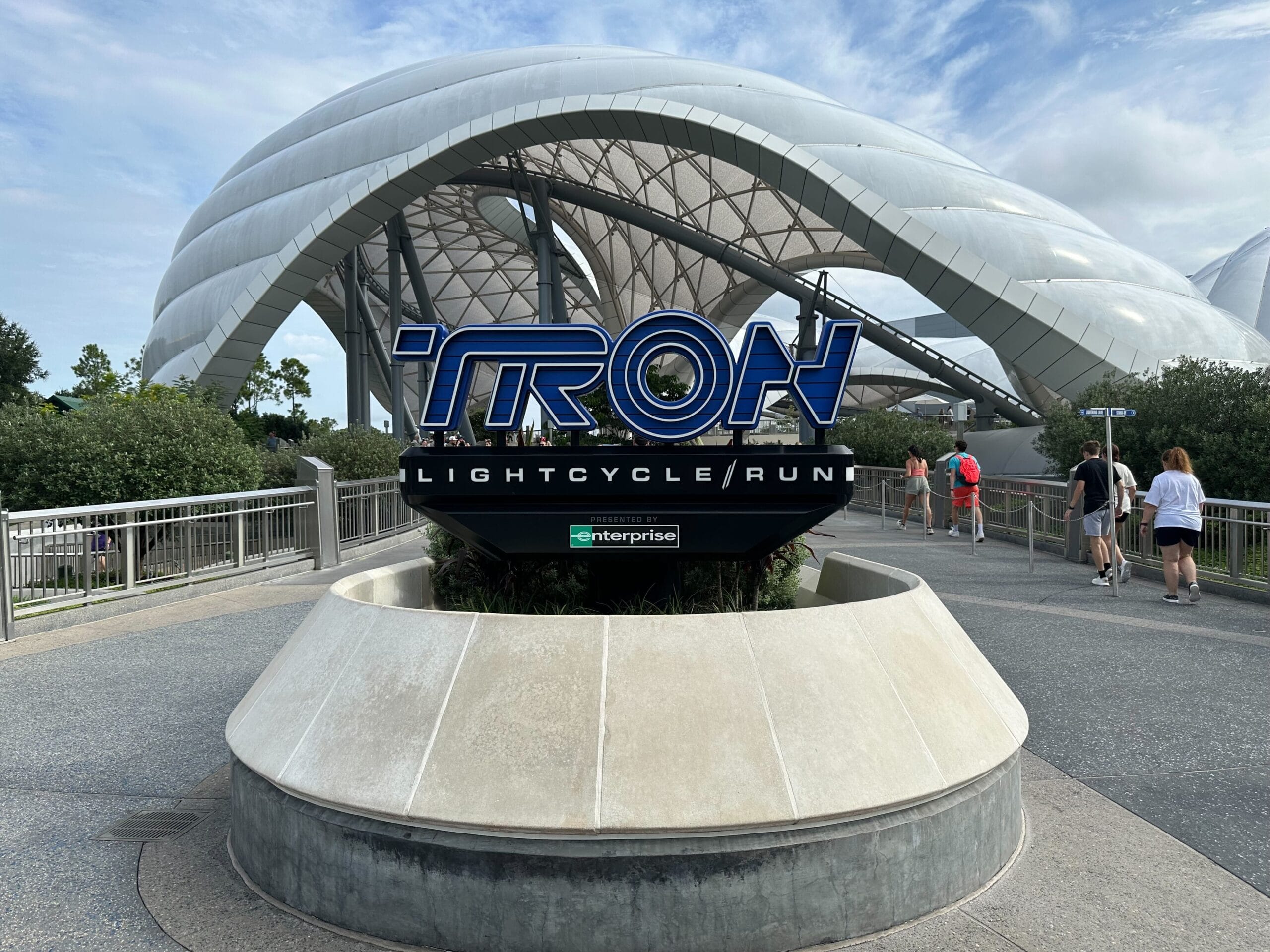 TRON Lightcycle / Run sign at Walt Disney World, set before a futuristic white canopy with visitors strolling nearby.