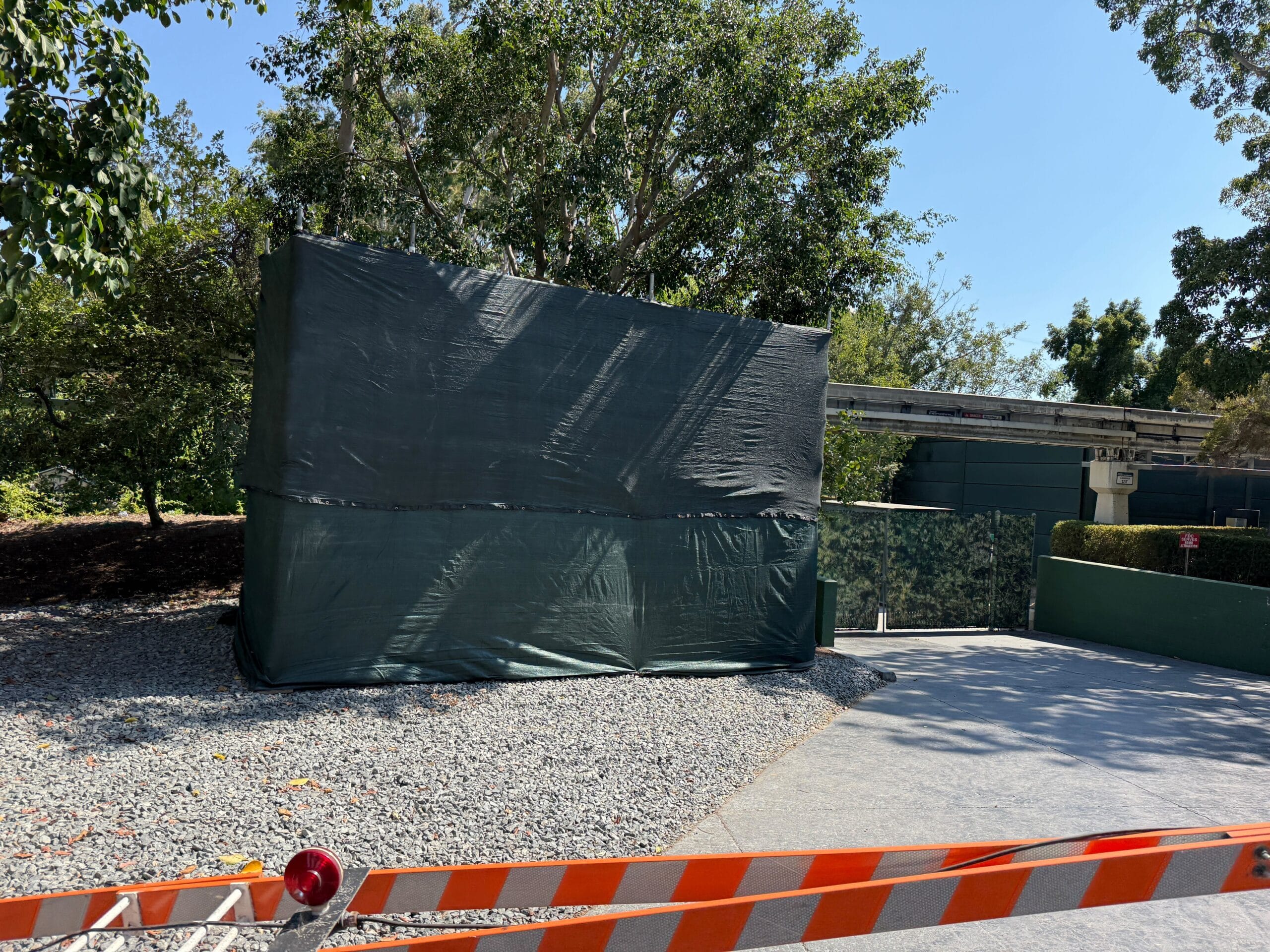 A large object covered in a dark tarp sits on gravel near trees, behind an orange-striped barrier on a sunny day.