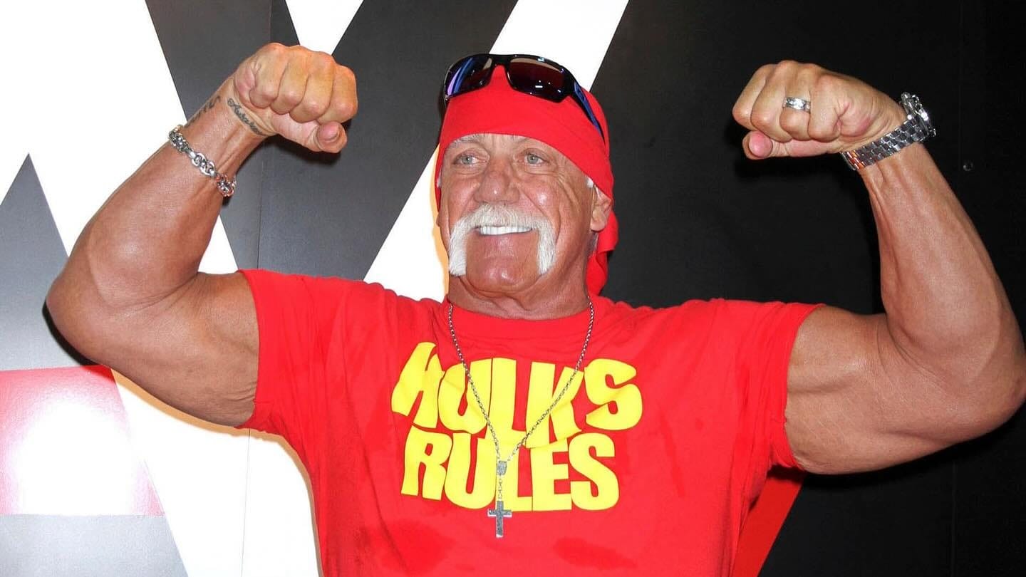 Muscular man in a red "Hulk's Rules" shirt flexes his arms and smiles, wearing a red bandana and sunglasses on his head.