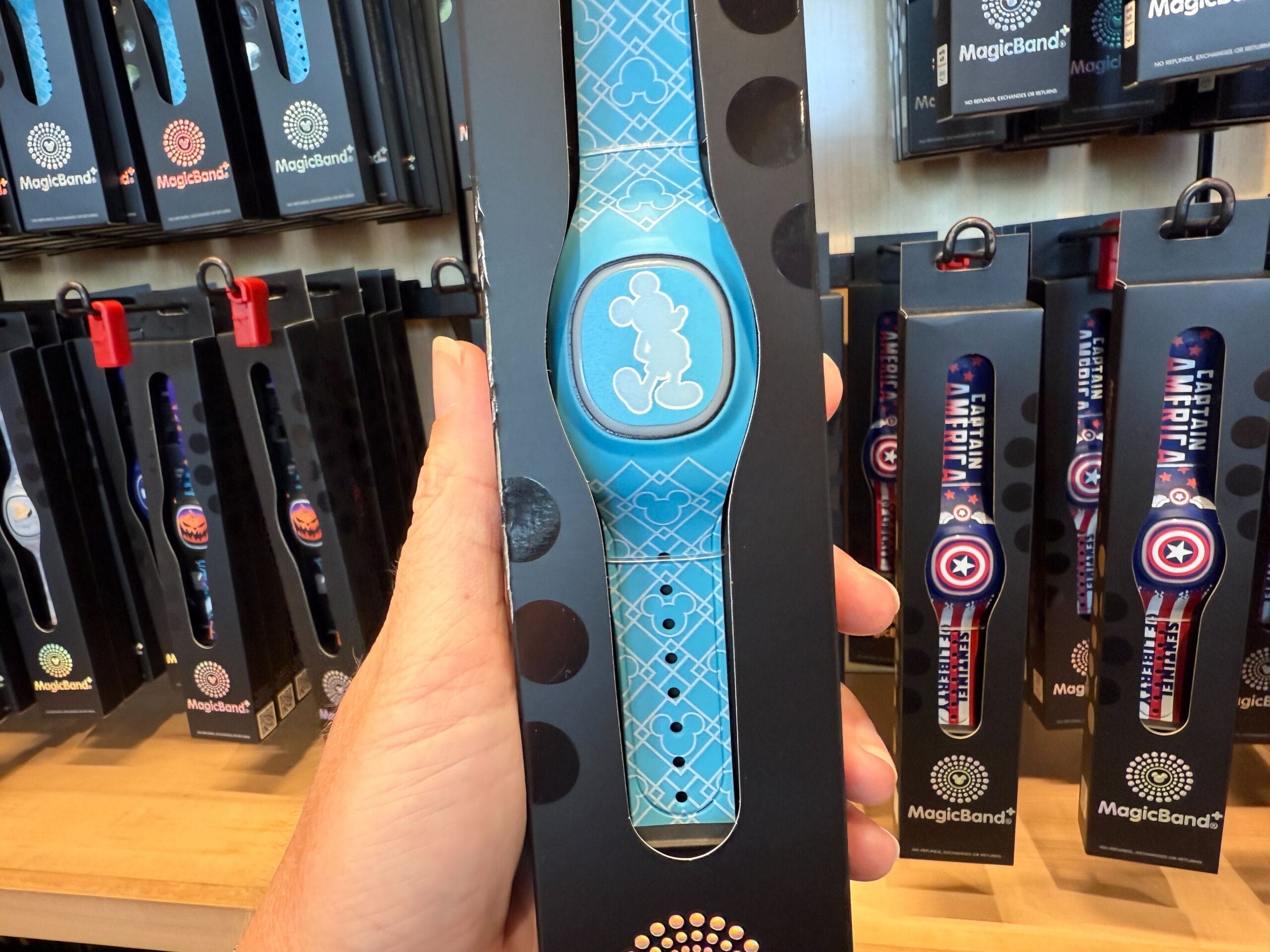 A hand holds a blue Disney MagicBand featuring a Mickey Mouse silhouette, with other bands displayed in the background.