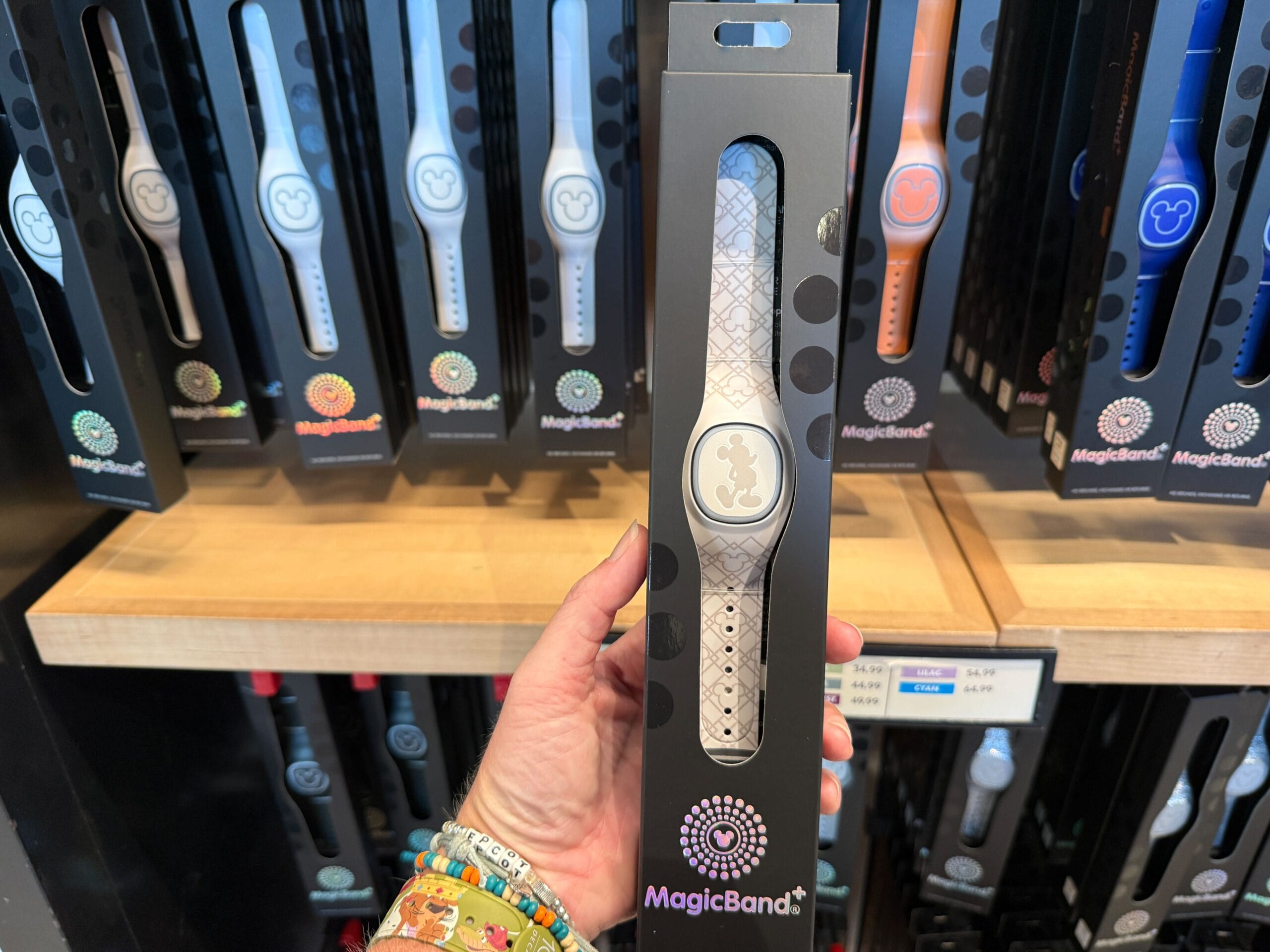 A hand holds a packaged MagicBand+ with a Mickey Mouse pattern in front of a display of assorted MagicBands.