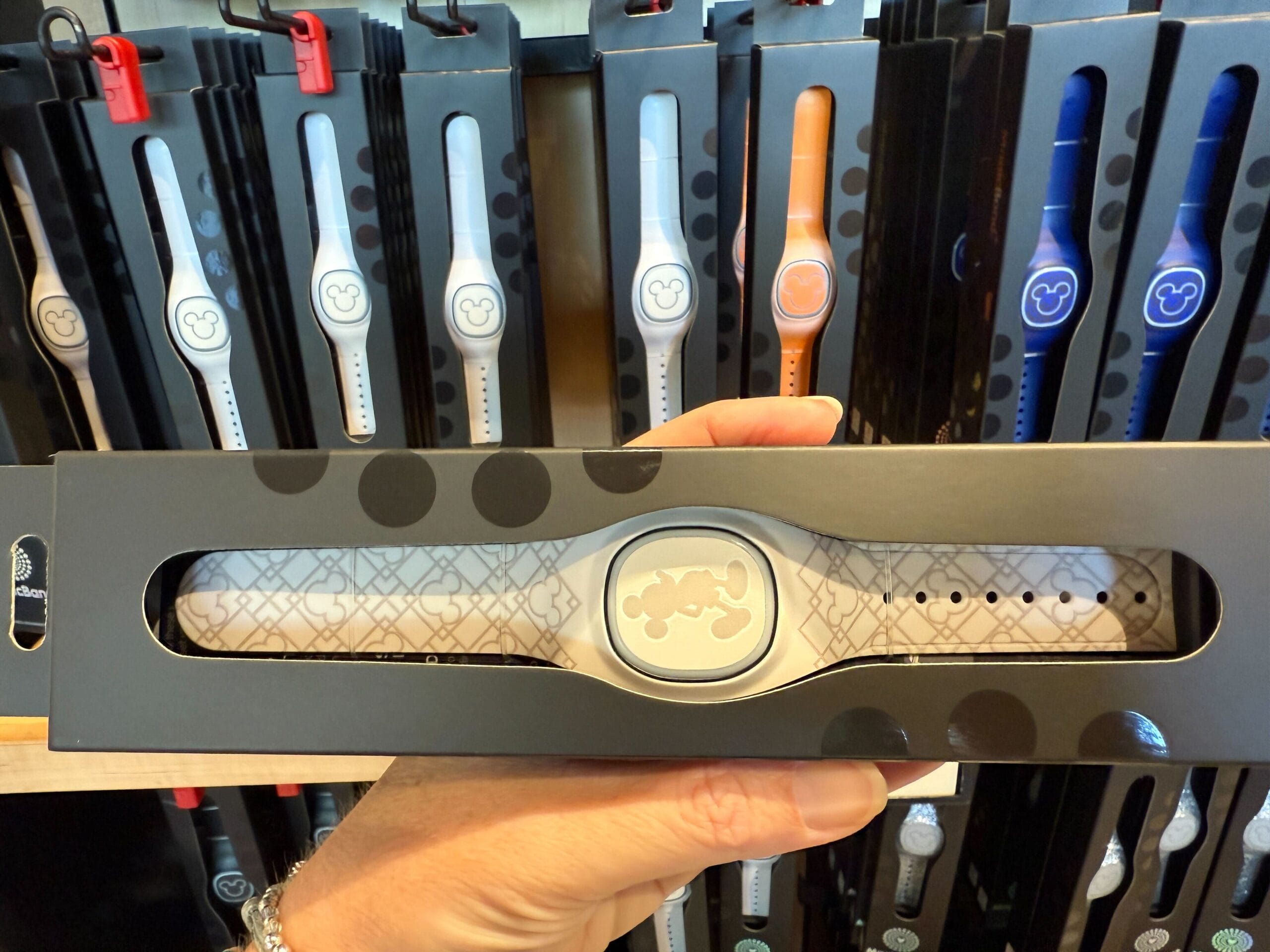 Hand holding a gray MagicBand+ with Mickey Mouse design; Art Deco-inspired MagicBands shown in the background.