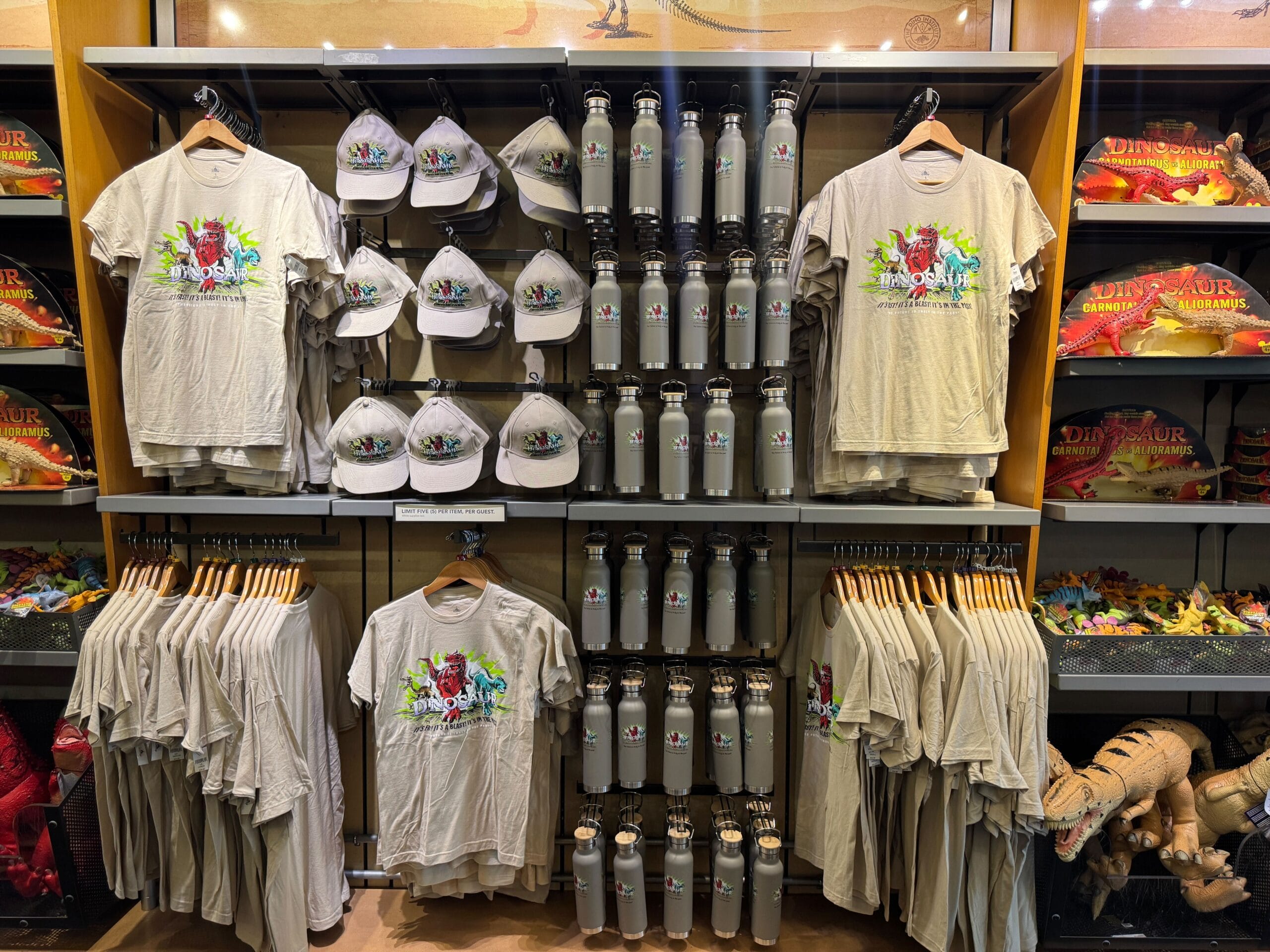 Dinosaur-themed t-shirts, hats, and water bottles displayed on shelves in a gift shop.