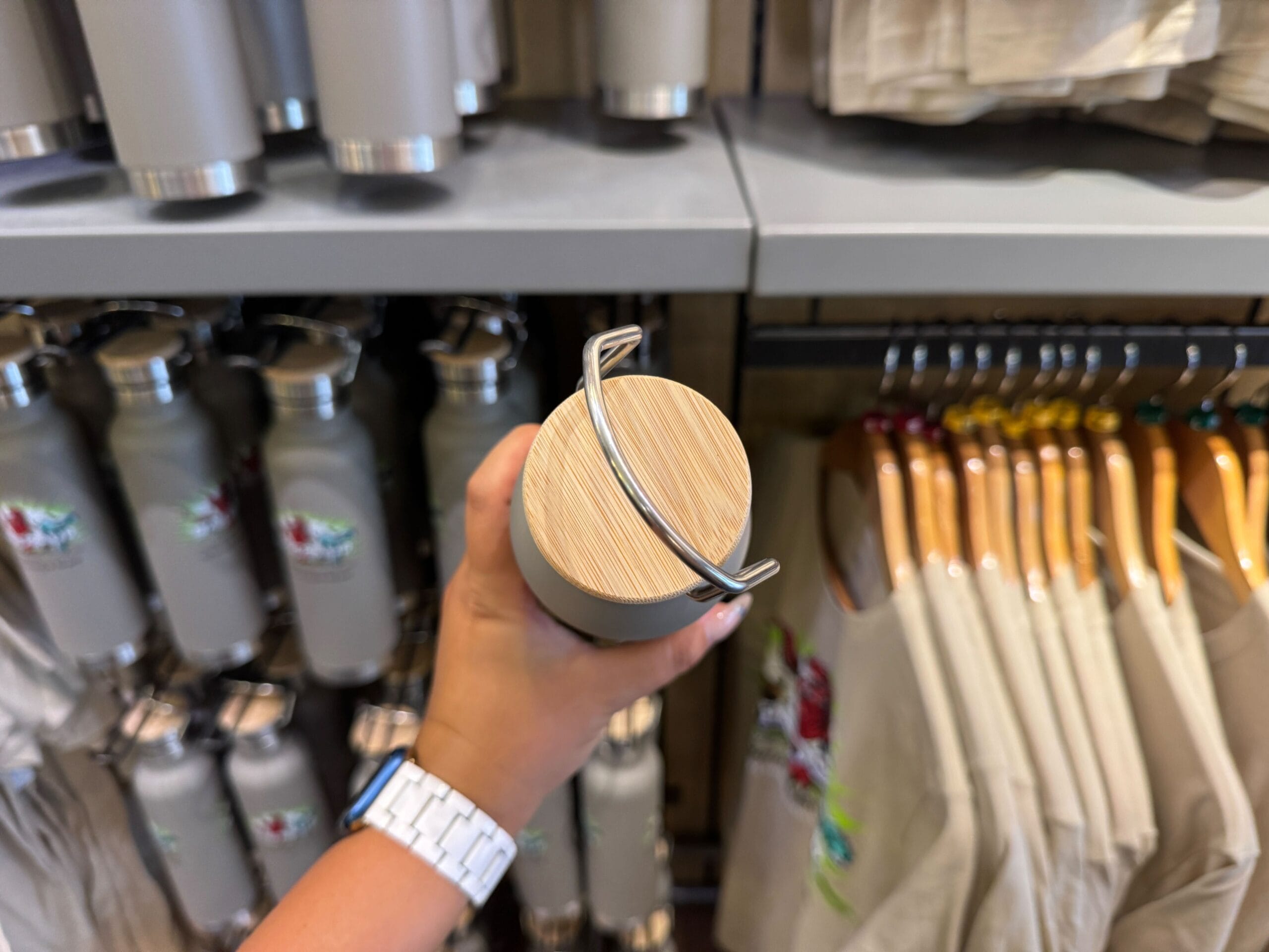 A hand holds a water bottle with a wooden lid in a store display of bottles and hanging shirts.