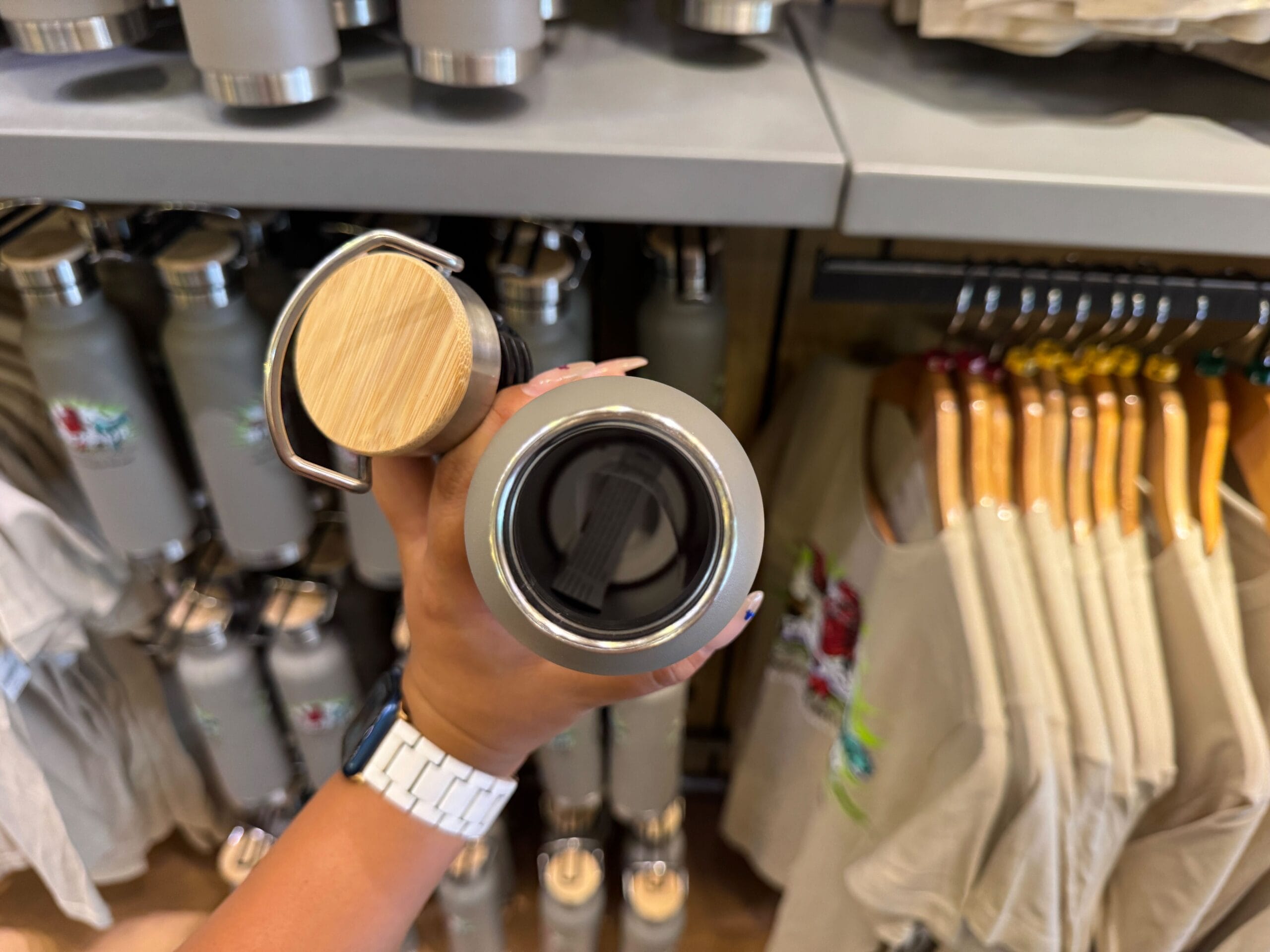 A hand holds an open metal water bottle with a wooden lid in a store selling bottles and clothes.