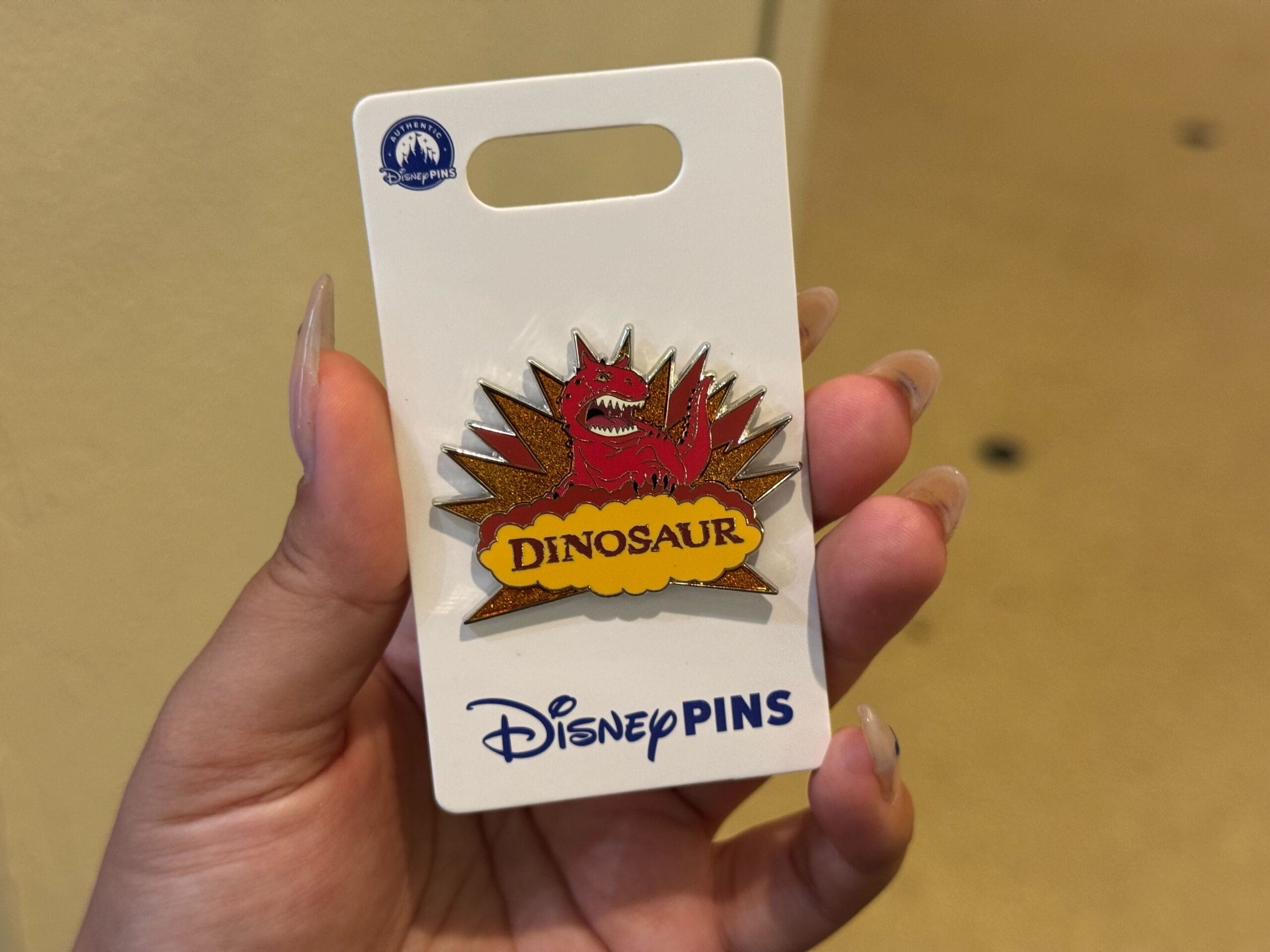 A hand holds a Disney pin featuring a red dinosaur and the word "DINOSAUR" on a starburst background.