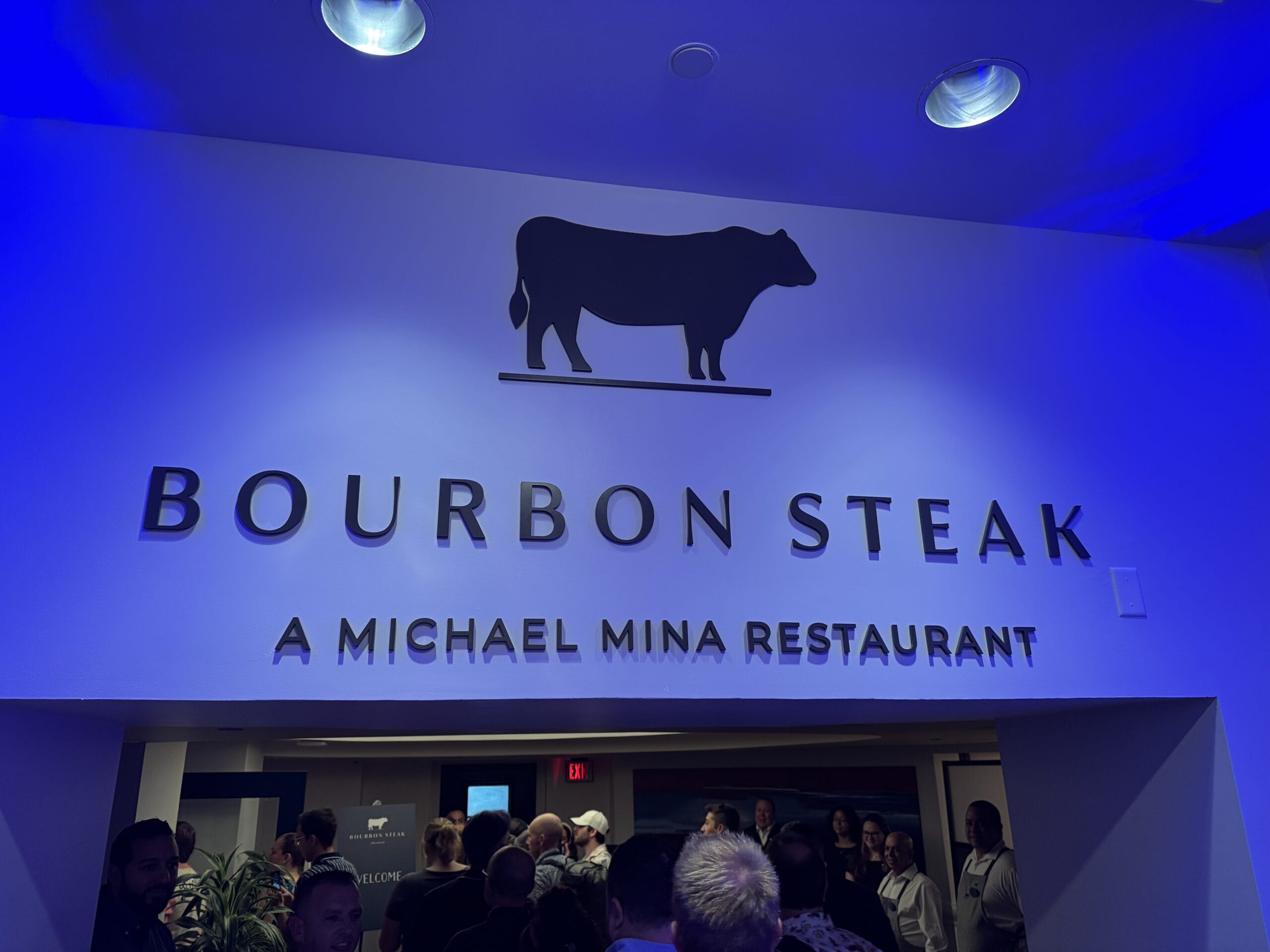 Entrance sign for Bourbon Steak by Michael Mina at Walt Disney World Dolphin Hotel, with people gathered below.