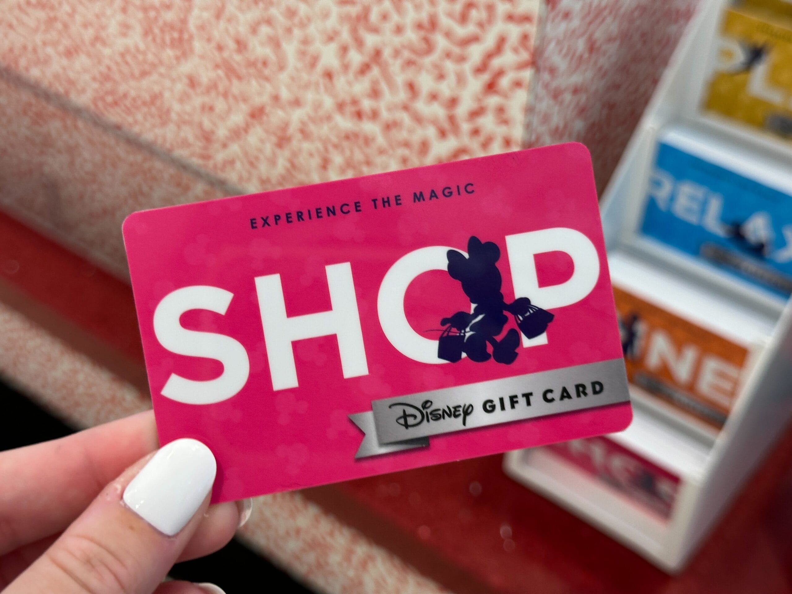Hand holding a pink Disney gift card that says "SHOP" with a silhouette of Mickey Mouse and the phrase "Experience the Magic.